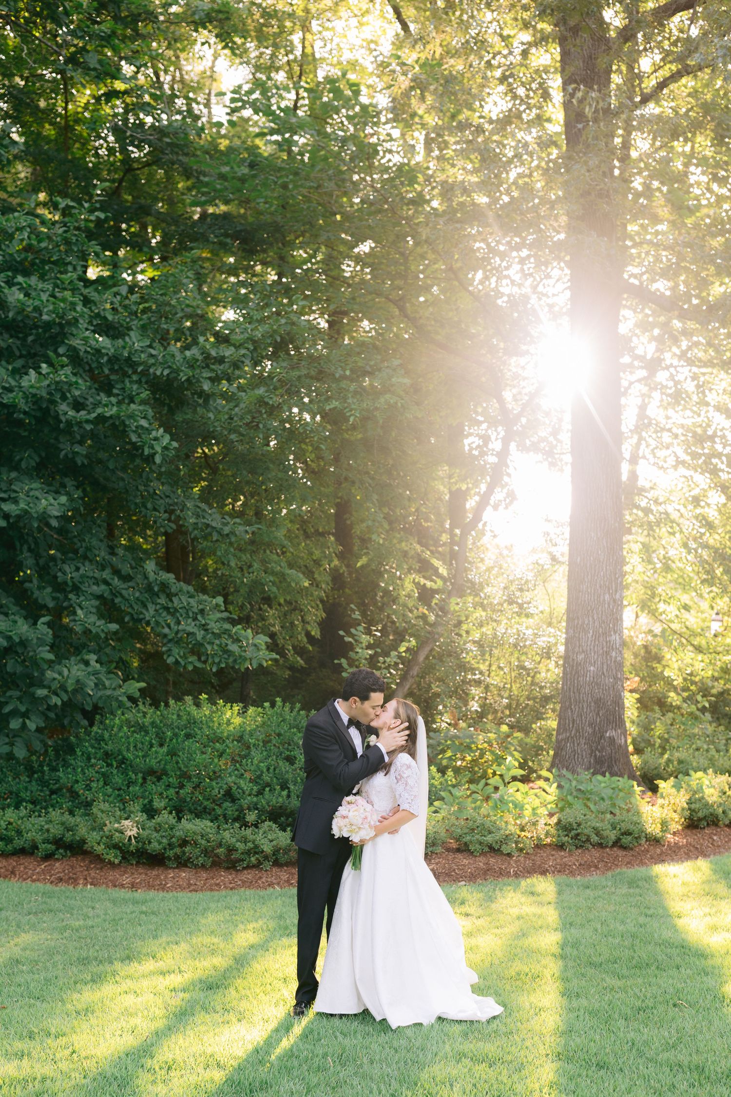 Wedding couple embraces in golden sunlight filtering through trees during sunset garden portraits.