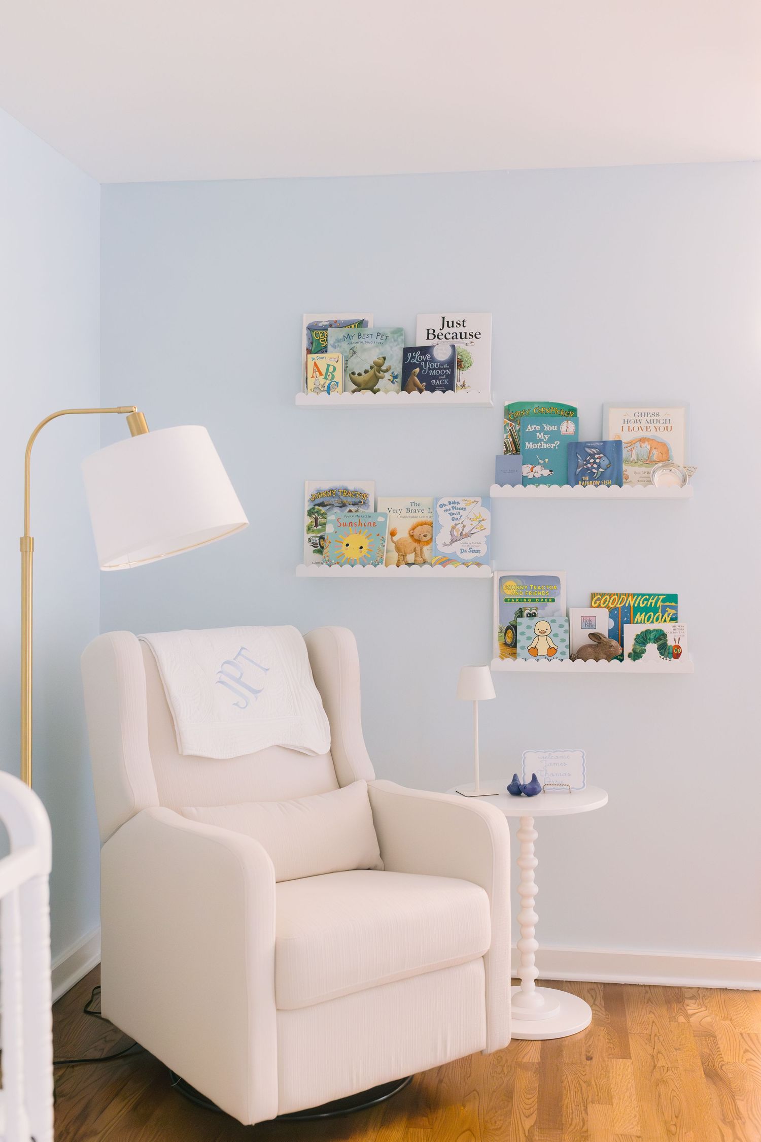 A cozy nursery corner features a white armchair, gold floor lamp, and floating shelves with children's books.