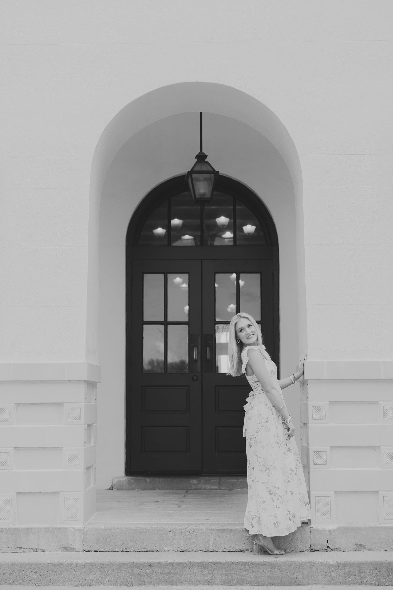Black and white artistic photography of person in white dress walking beside white building with arched doorways.