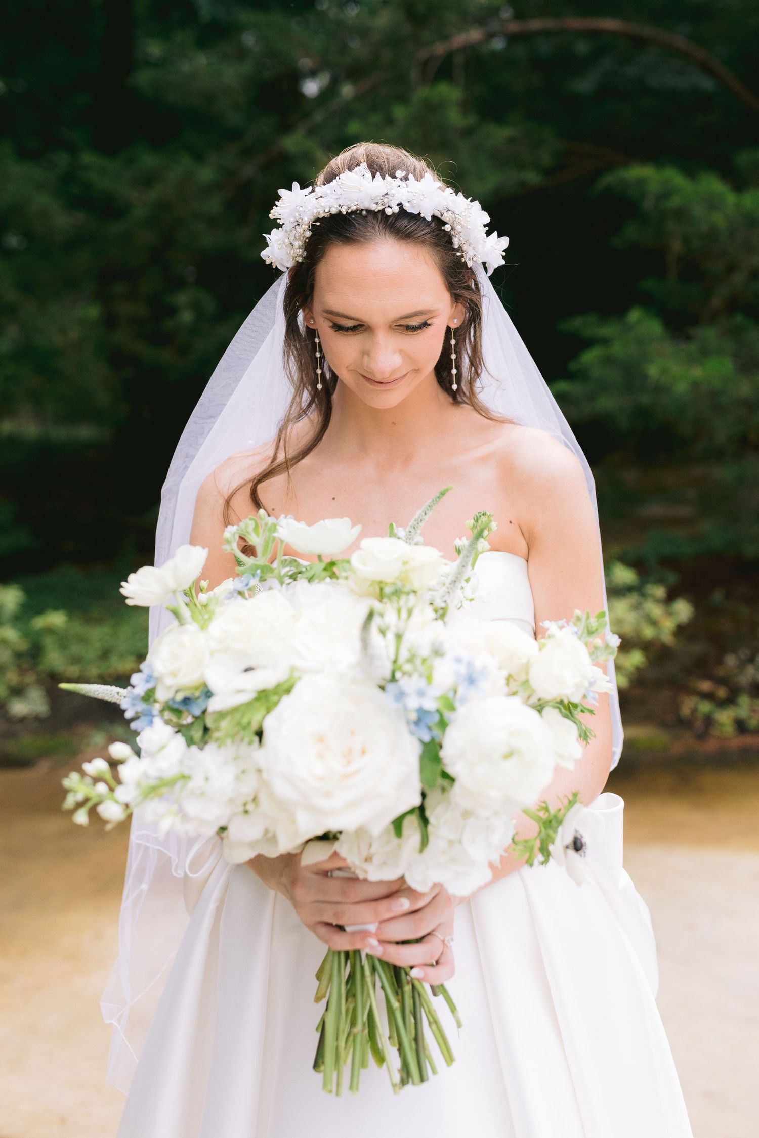 A bride in a flowing white dress holds an elegant bouquet of white flowers and delicate greenery at an outdoor ceremony.