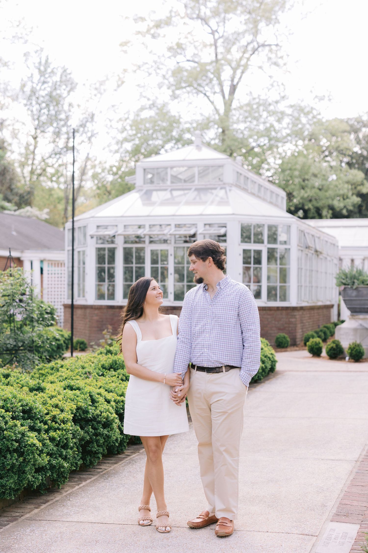 Two people stand together on a pathway near white greenhouse buildings surrounded by manicured shrubs and landscaping.