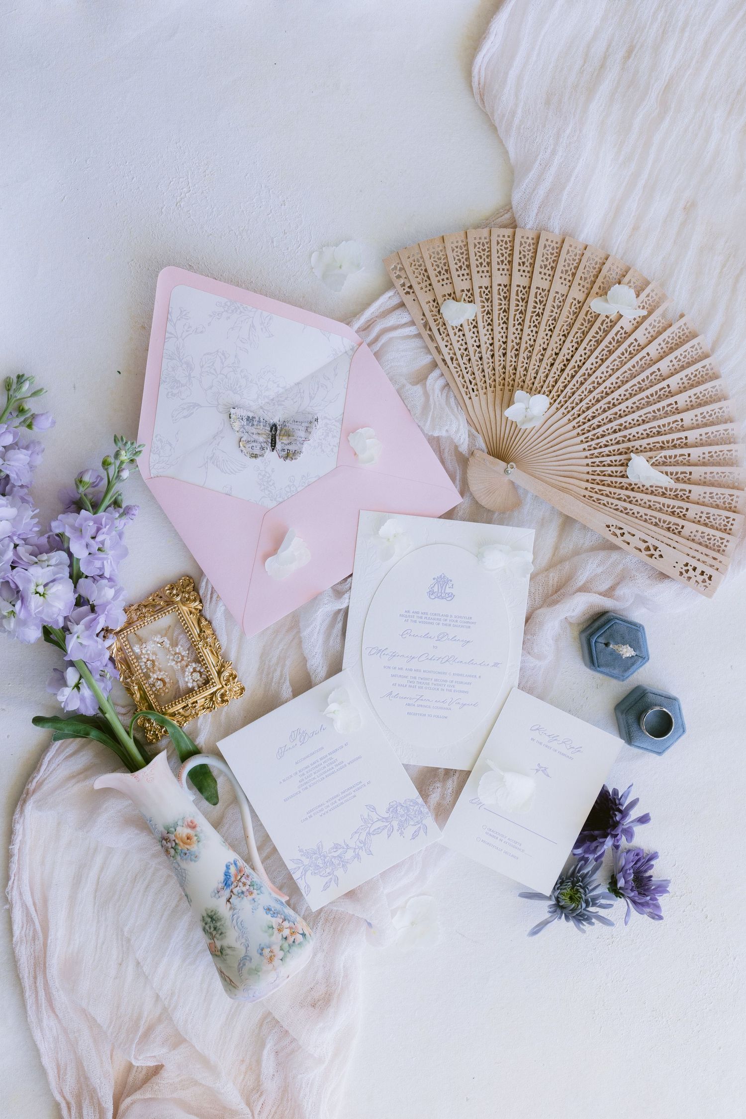 Elegant wedding details featuring paper invitations, decorative fans, and purple flowers arranged on white fabric.