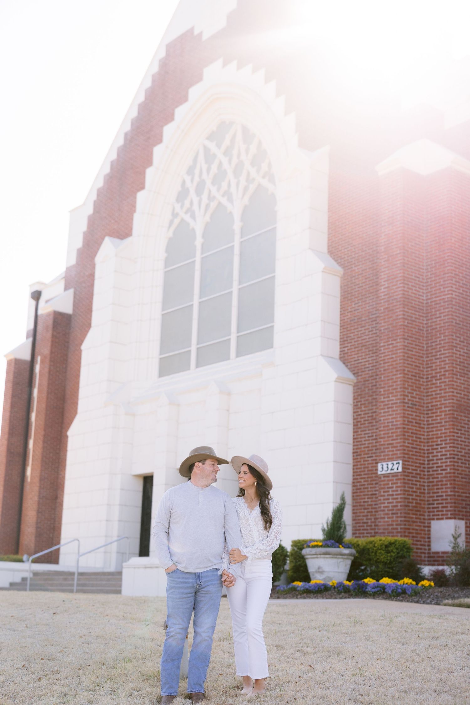 A couple stands together outside a brick church with a white arched entrance and yellow flowers near the doorway.