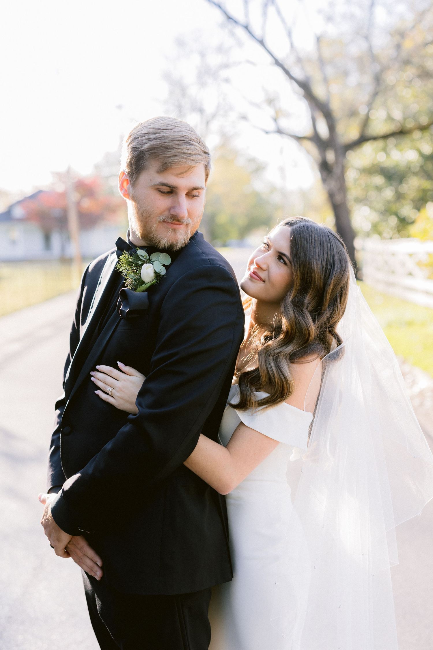 Bride in white dress and groom in black suit embrace outdoors on their wedding day with autumn trees in background.