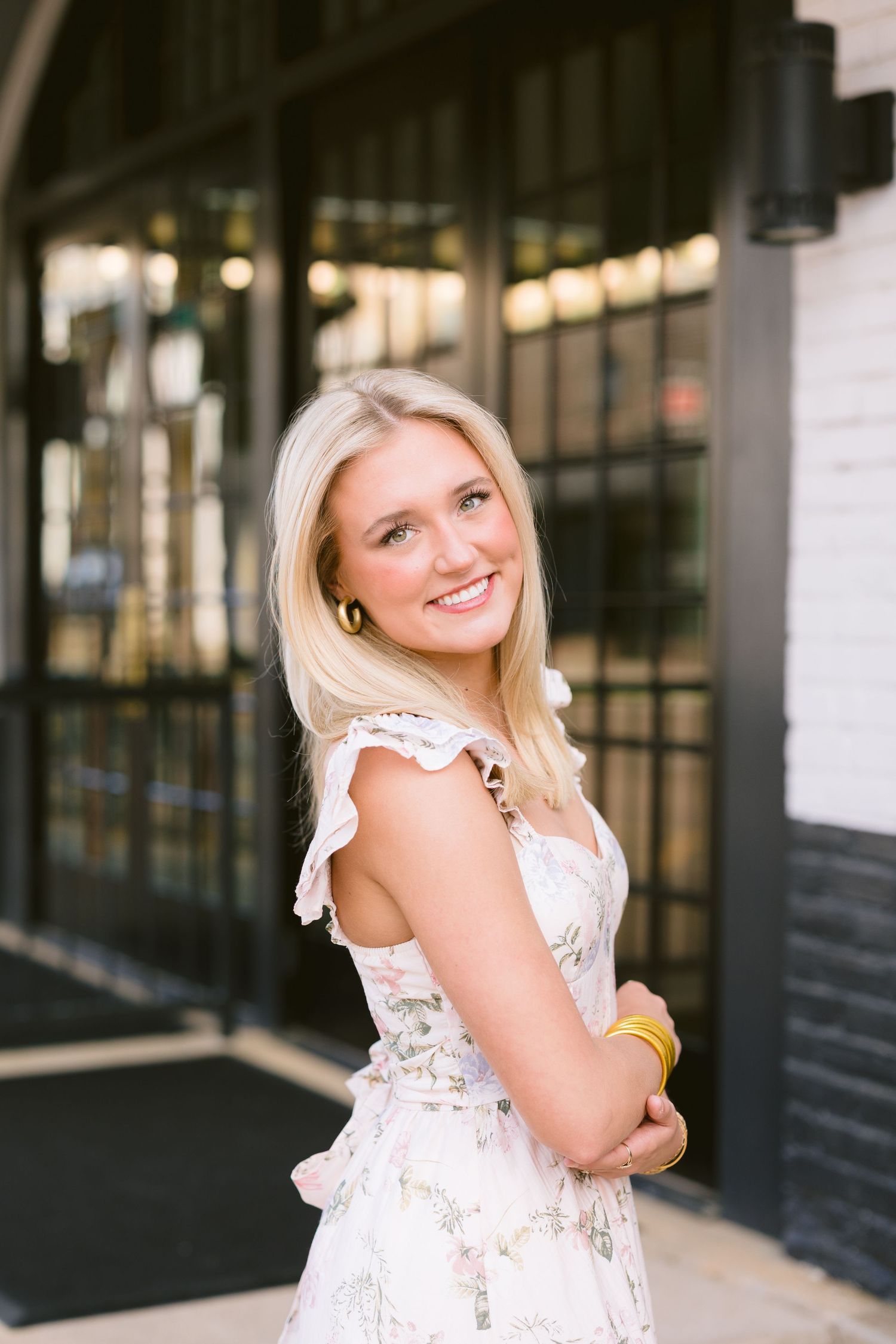 A person in a white lace dress with ruffled sleeves poses outdoors near a storefront with string lights.