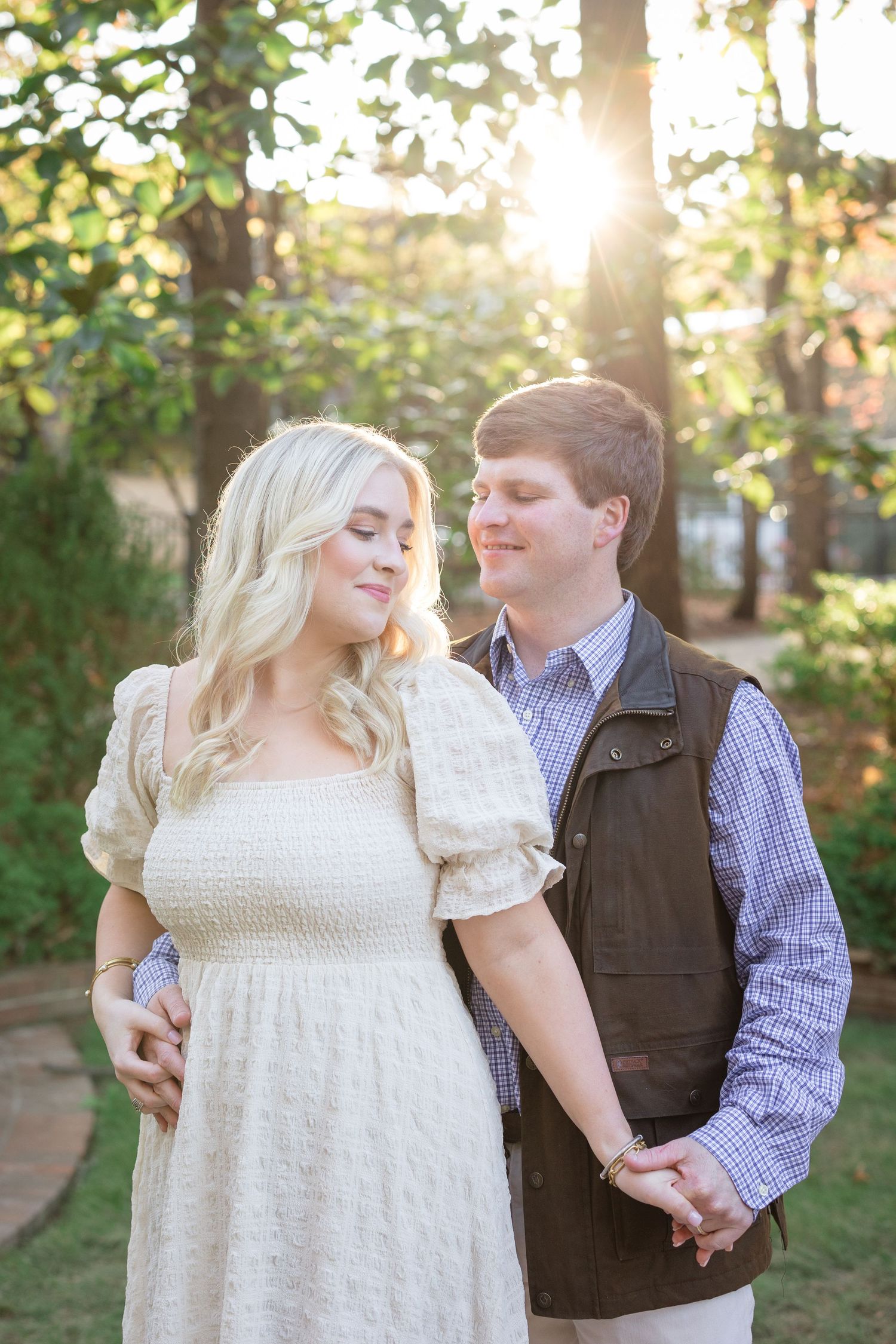 A couple shares a romantic moment outdoors during golden hour among trees wearing a white dress and brown vest.