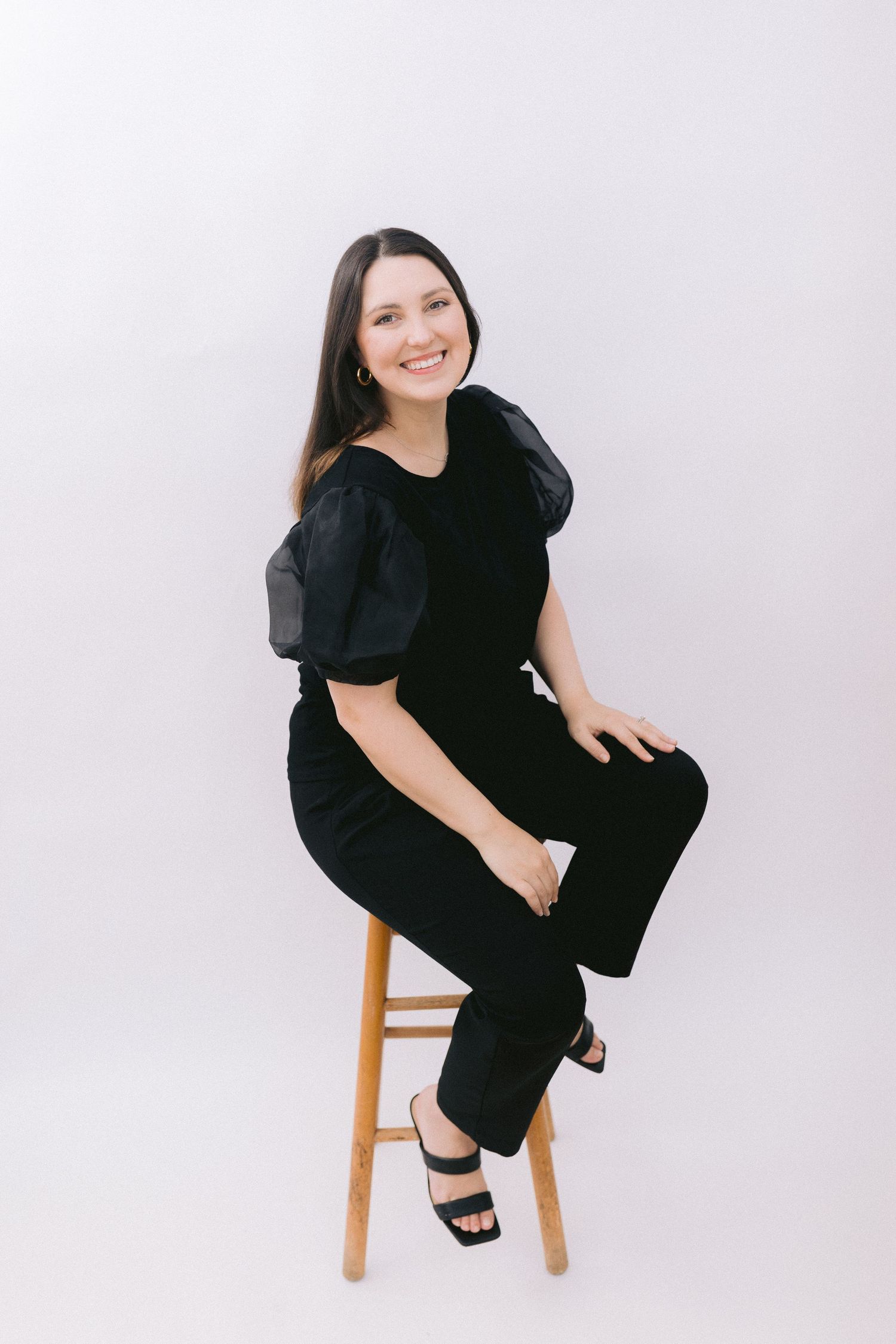 Woman in black outfit poses confidently on wooden stool against white backdrop.