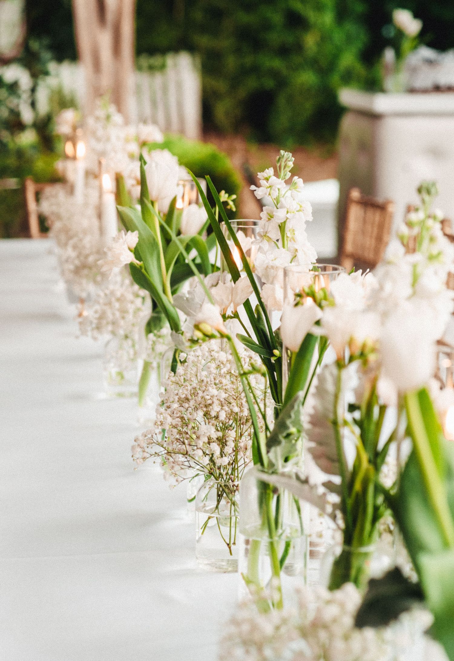 White floral centerpieces with delicate baby's breath in glass vases lined up on reception table.