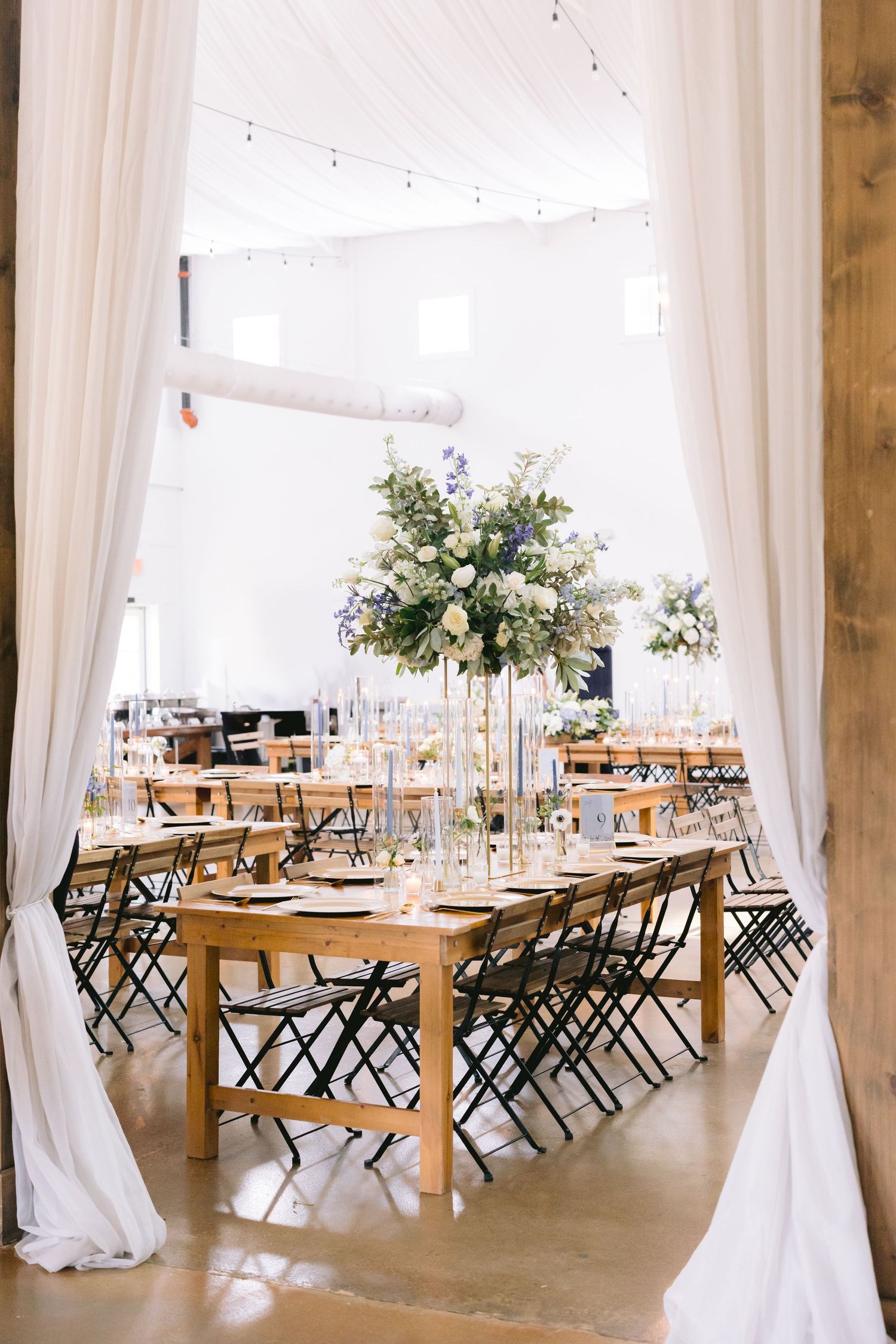 Elegant wedding reception venue with white drapes, wooden tables and cross-back chairs.