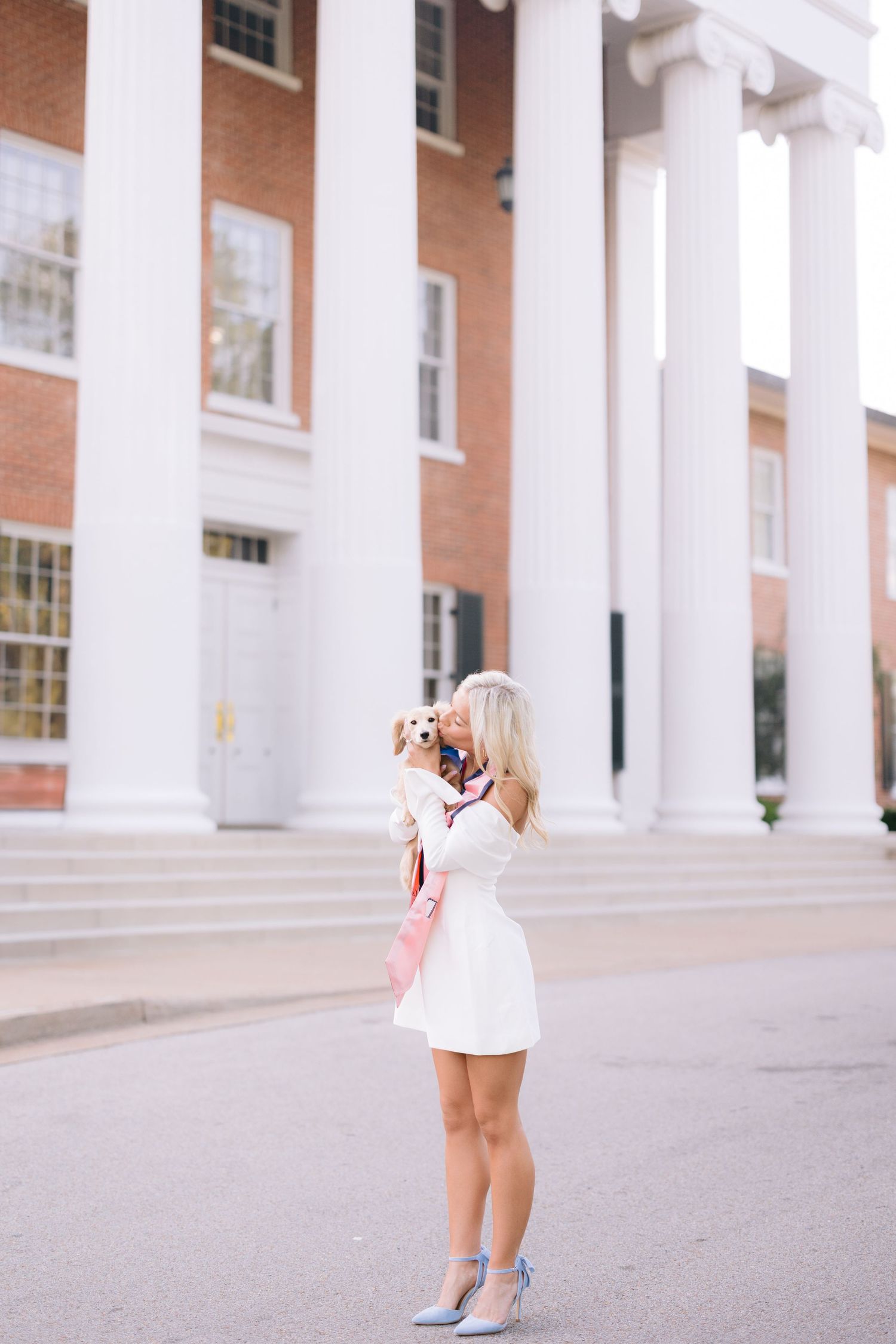 Figure in a white dress holding flowers stands in front of a brick building with large white columns in elegant pose.