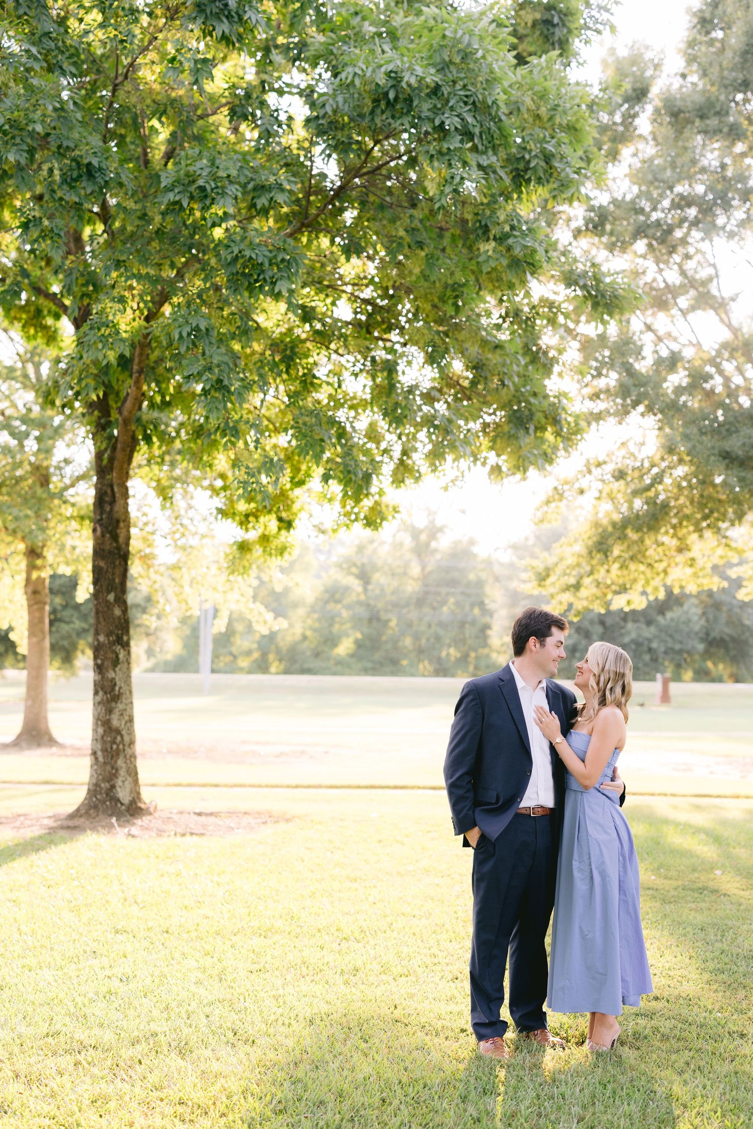 A couple shares tender moments in a serene park setting with sunlit trees during a romantic outdoor photo session.