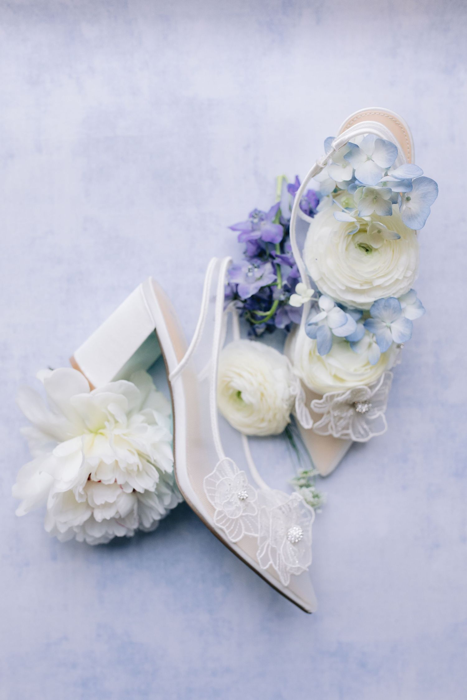 Delicate white and blue floral arrangements displayed alongside elegant bridal shoes on a soft gray background.