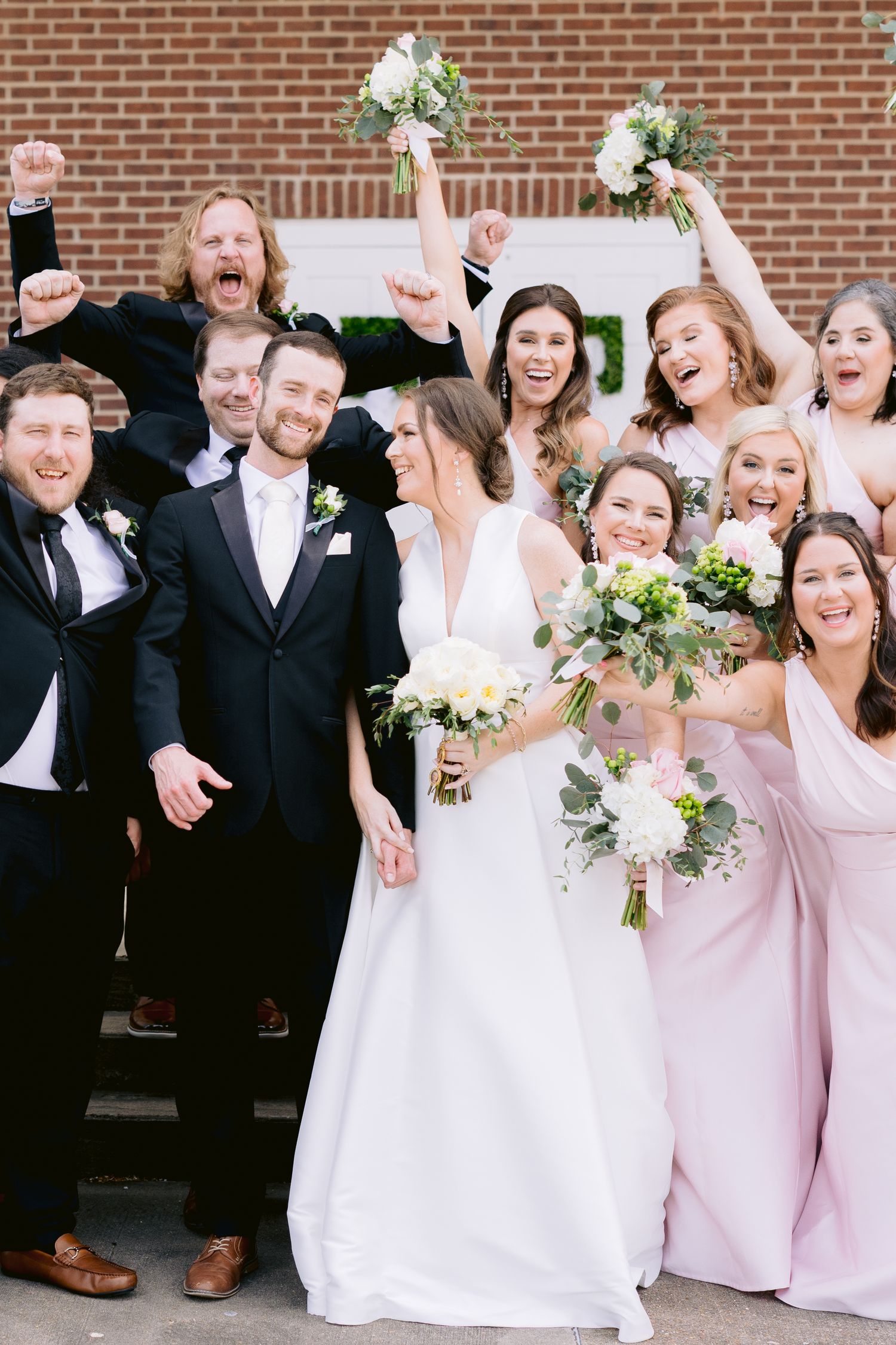 Wedding party celebrating with joyful expressions in front of brick wall.