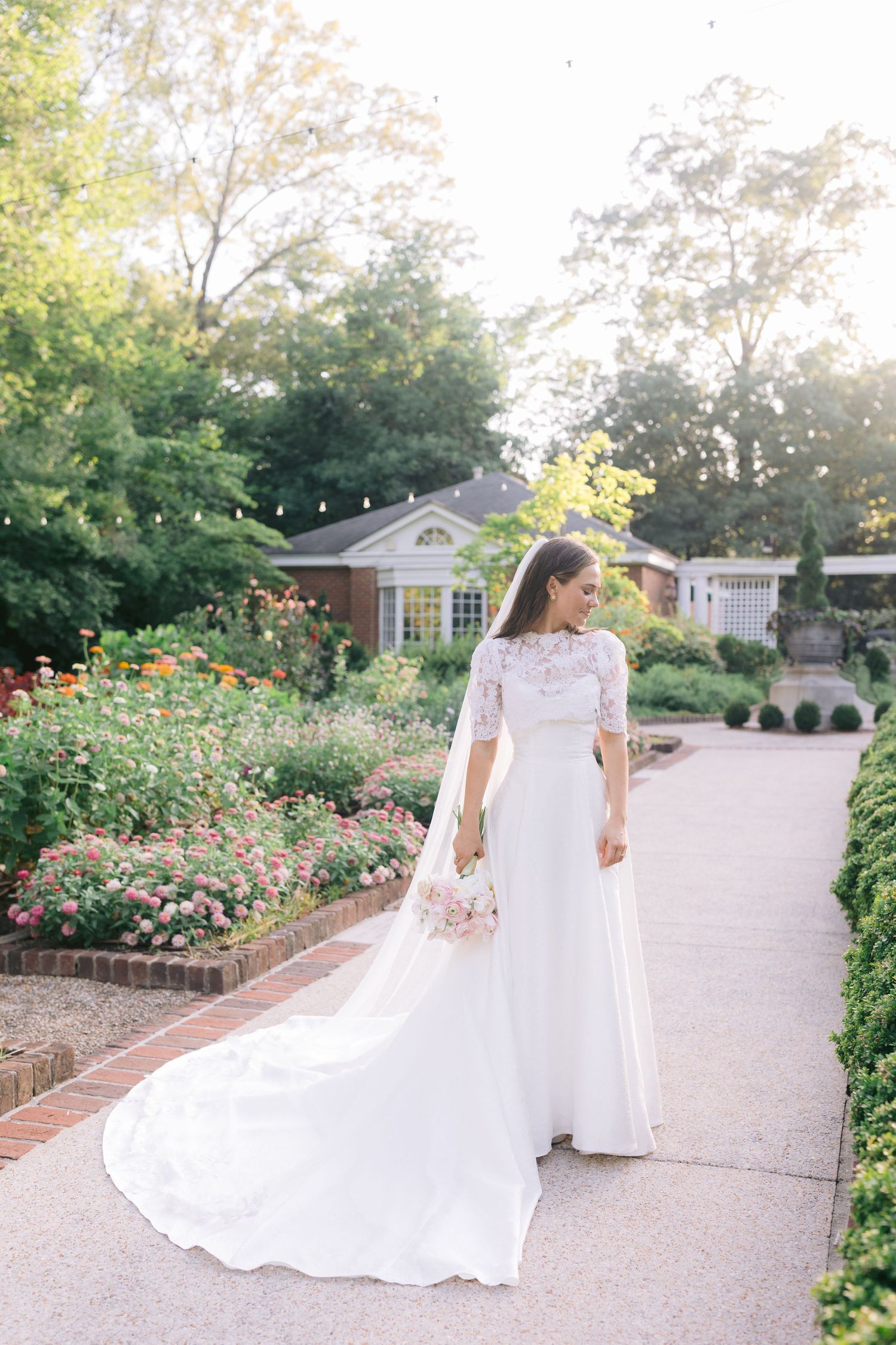Elegant bride in a white gown walks through a garden path lined with vibrant roses.
