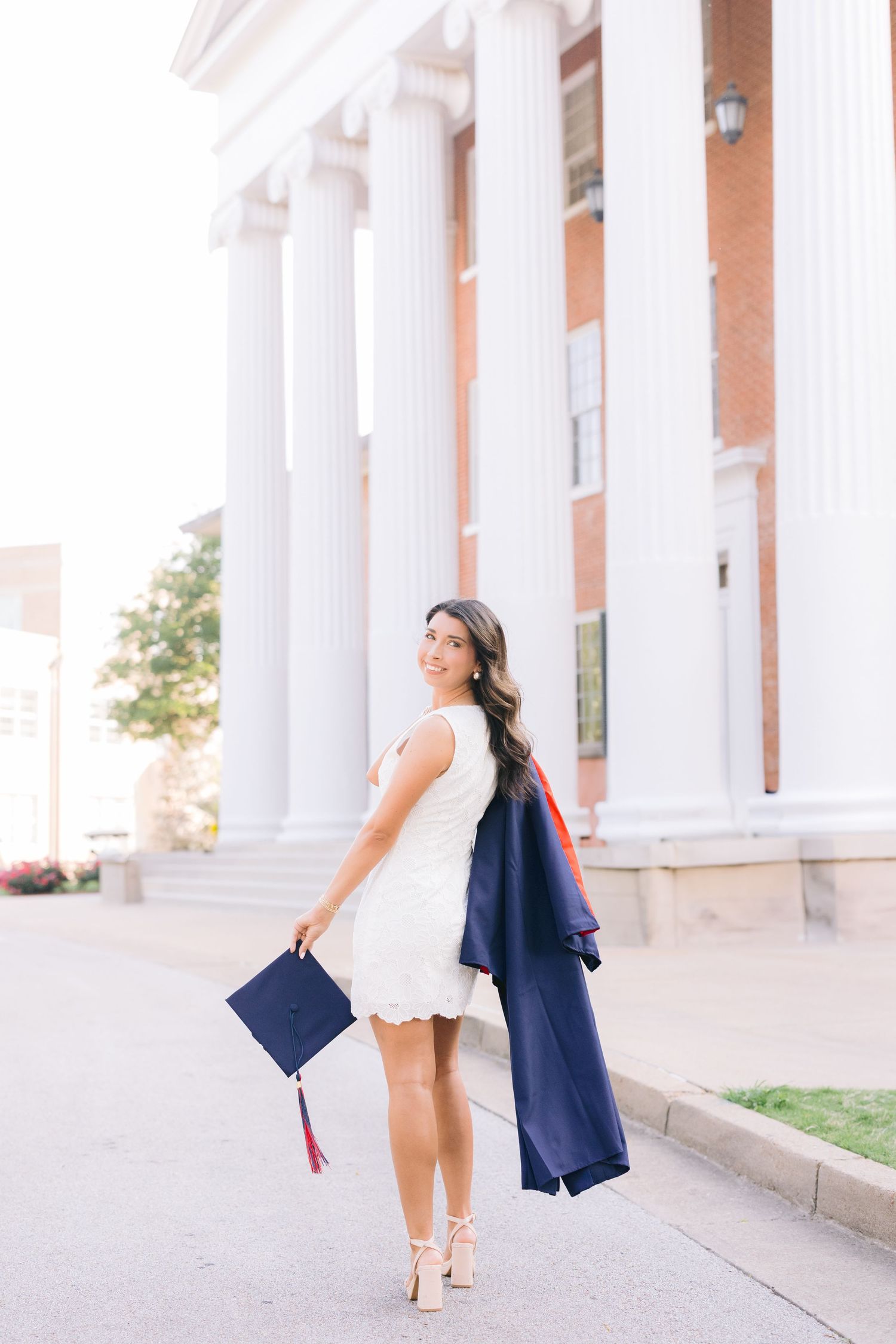 Graduate throws graduation cap and navy gown while standing by white pillars in elegant white dress and heels.