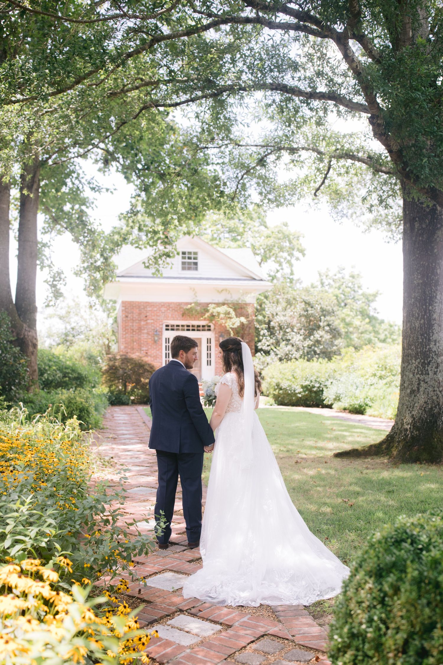 A wedding couple walks along a brick pathway surrounded by lush gardens and trees near a vintage brick building at Hedge Farms.