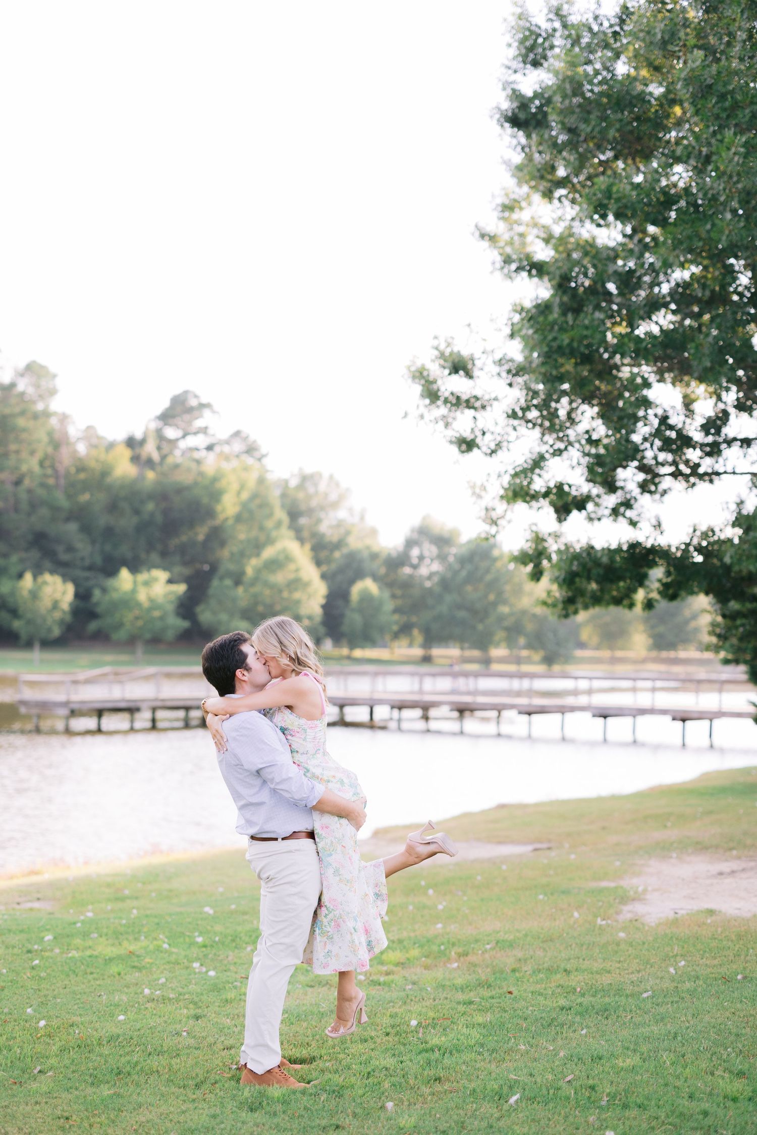 A couple in white outfits share a romantic embrace near a lake and fence at sunset.