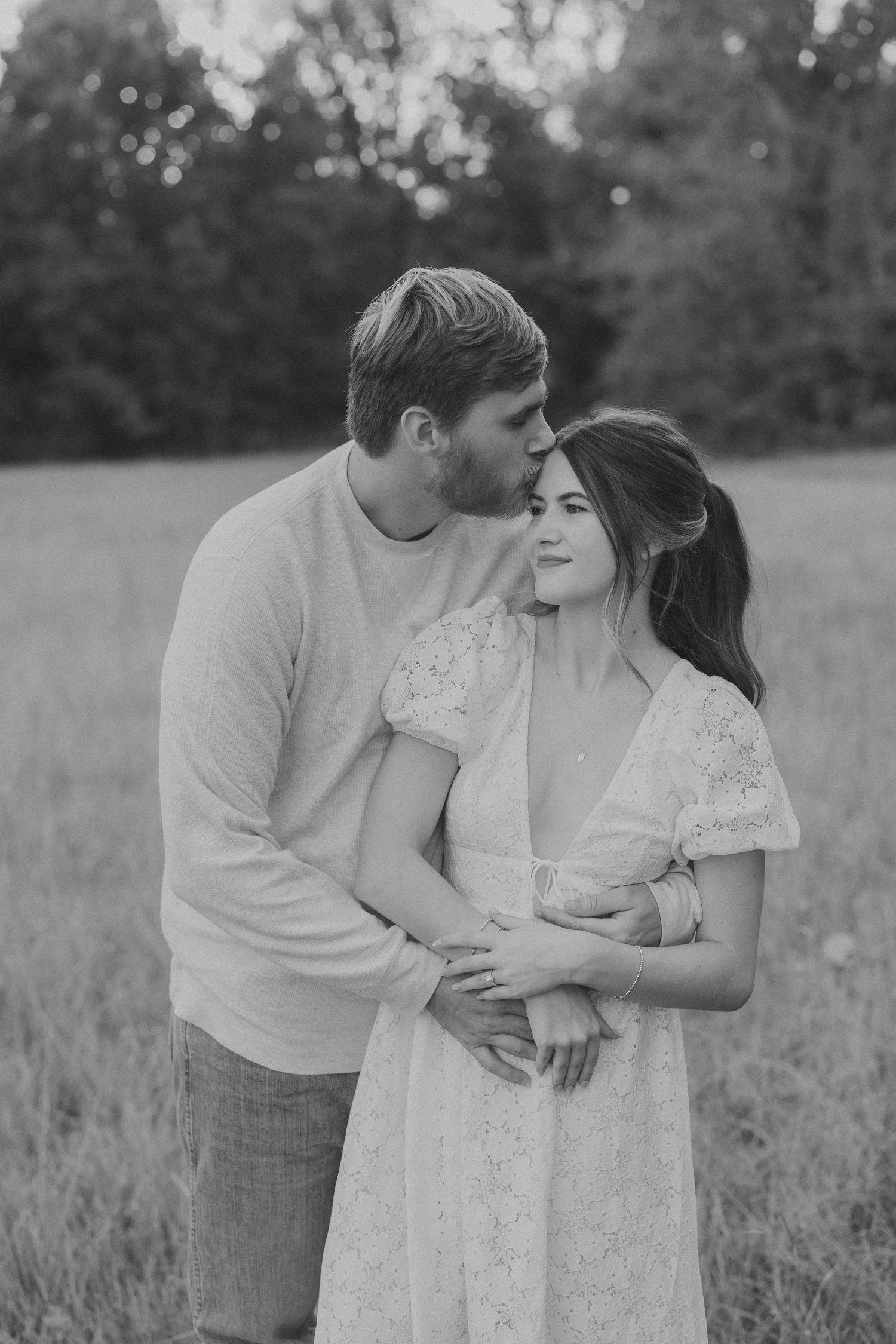 A romantic couple embraces in a serene meadow field wearing casual white attire in black and white photography.