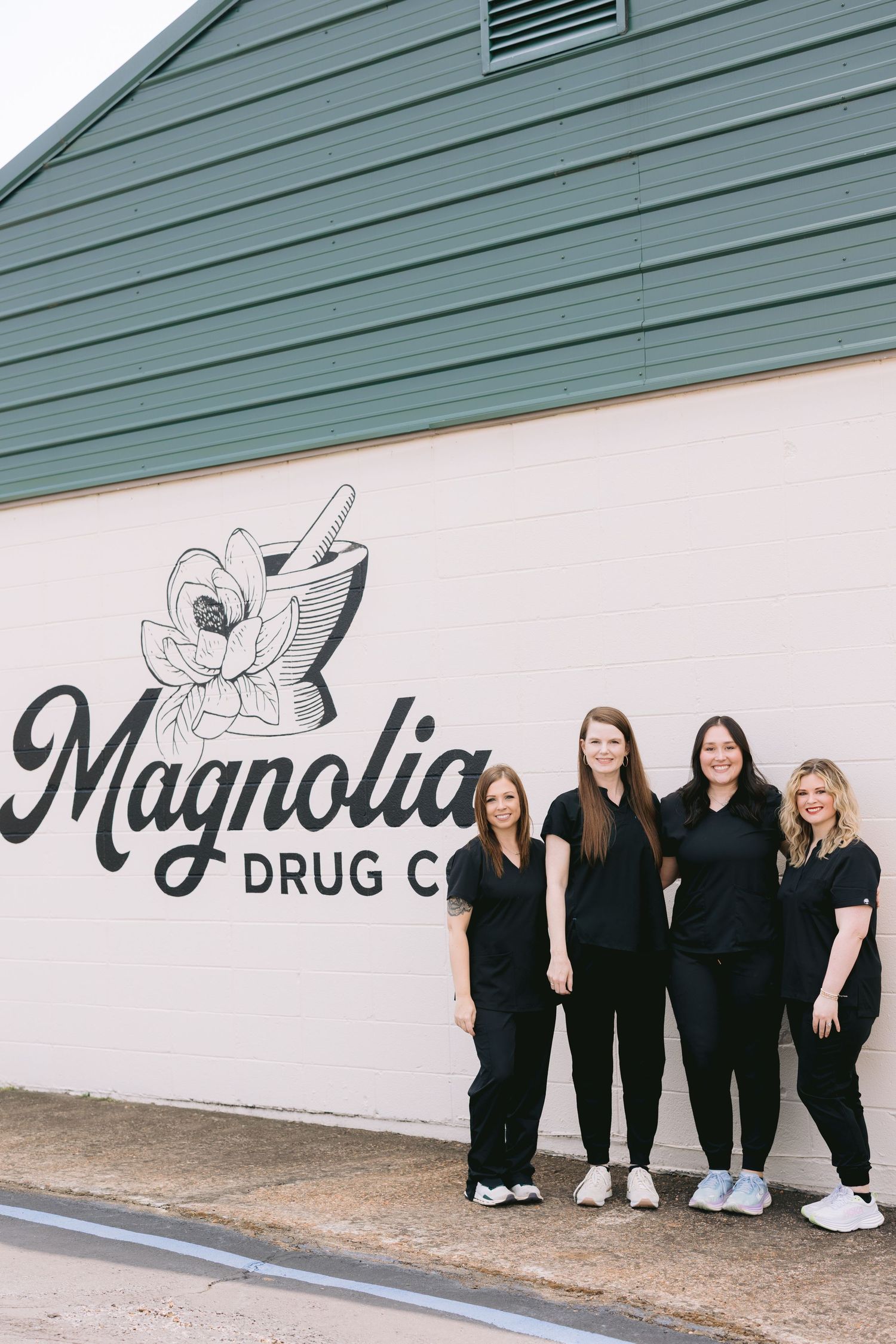 Group wearing black outfits poses casually against white wall with Magnolia Drug Co signage.