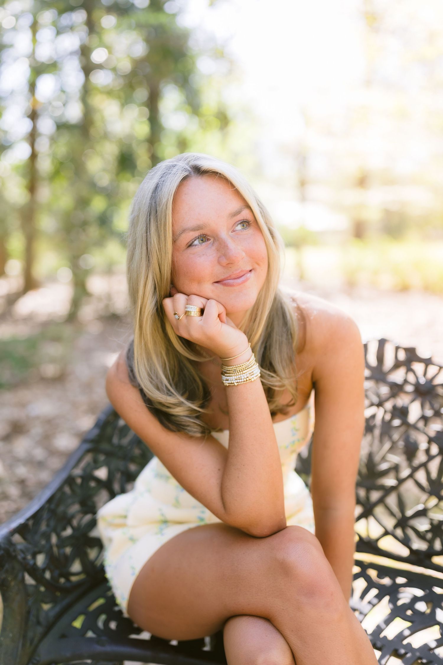 Smiling blonde model poses outdoors in natural setting while resting on decorative iron bench.