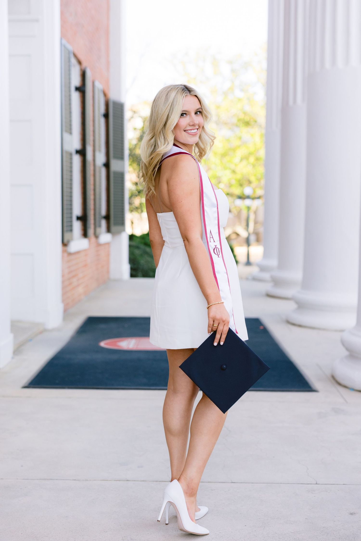 A graduate poses in a white dress and heels on a columned porch, holding a black diploma portfolio for portraits.