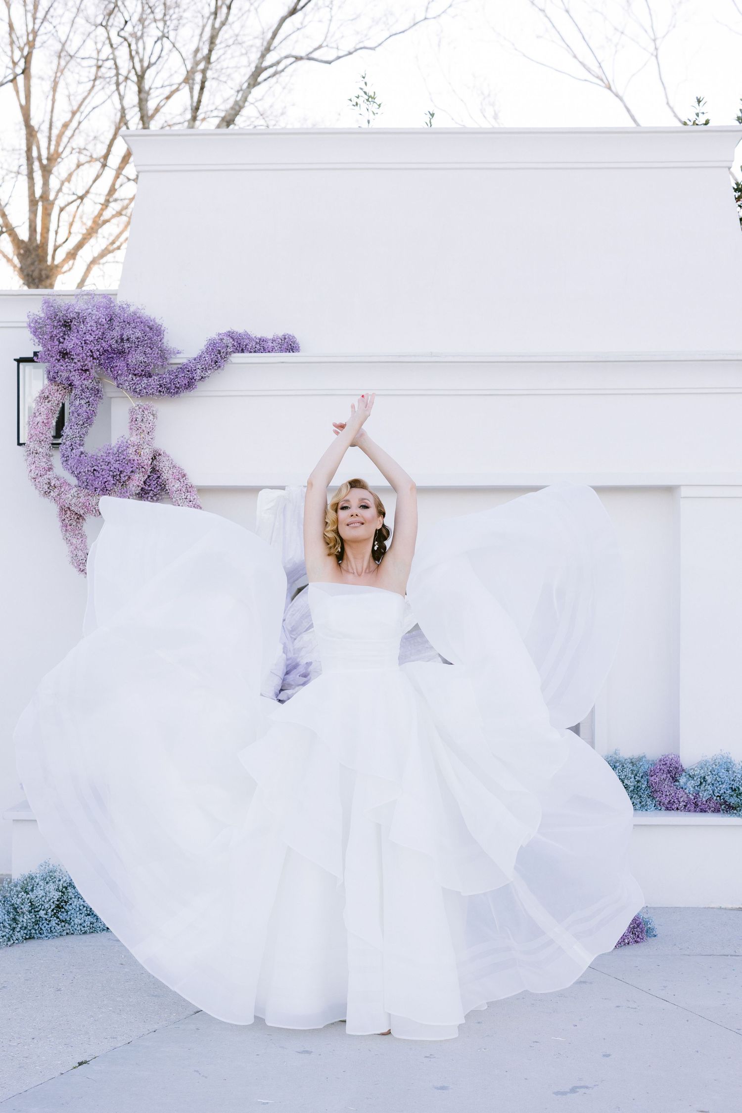 Ethereal white wedding dress with flowing layers creates dreamy movement against a backdrop with purple flowers.