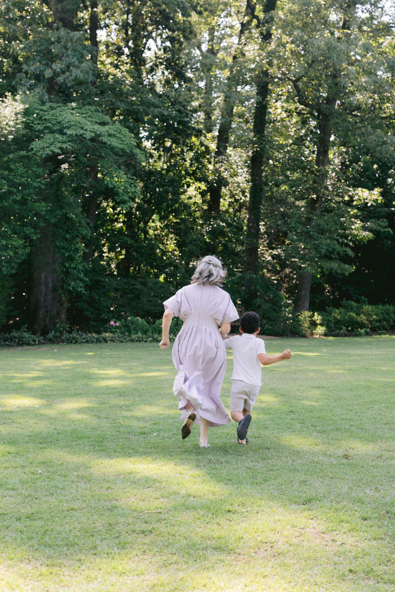 Two figures in light colored clothing run playfully across a sunlit grassy lawn bordered by trees.