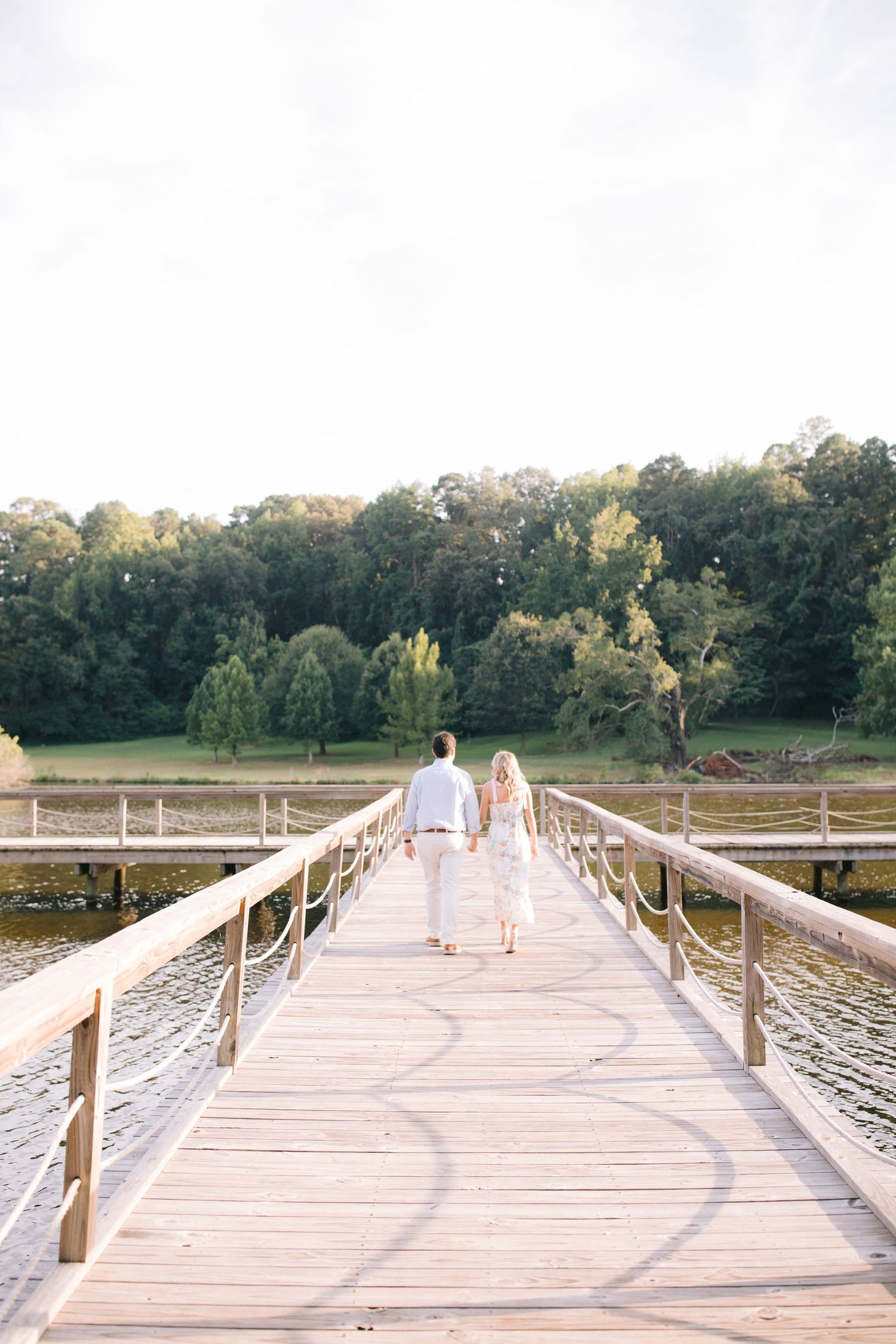 Couple walking hand in hand across a long wooden boardwalk over water at sunset with trees in background.
