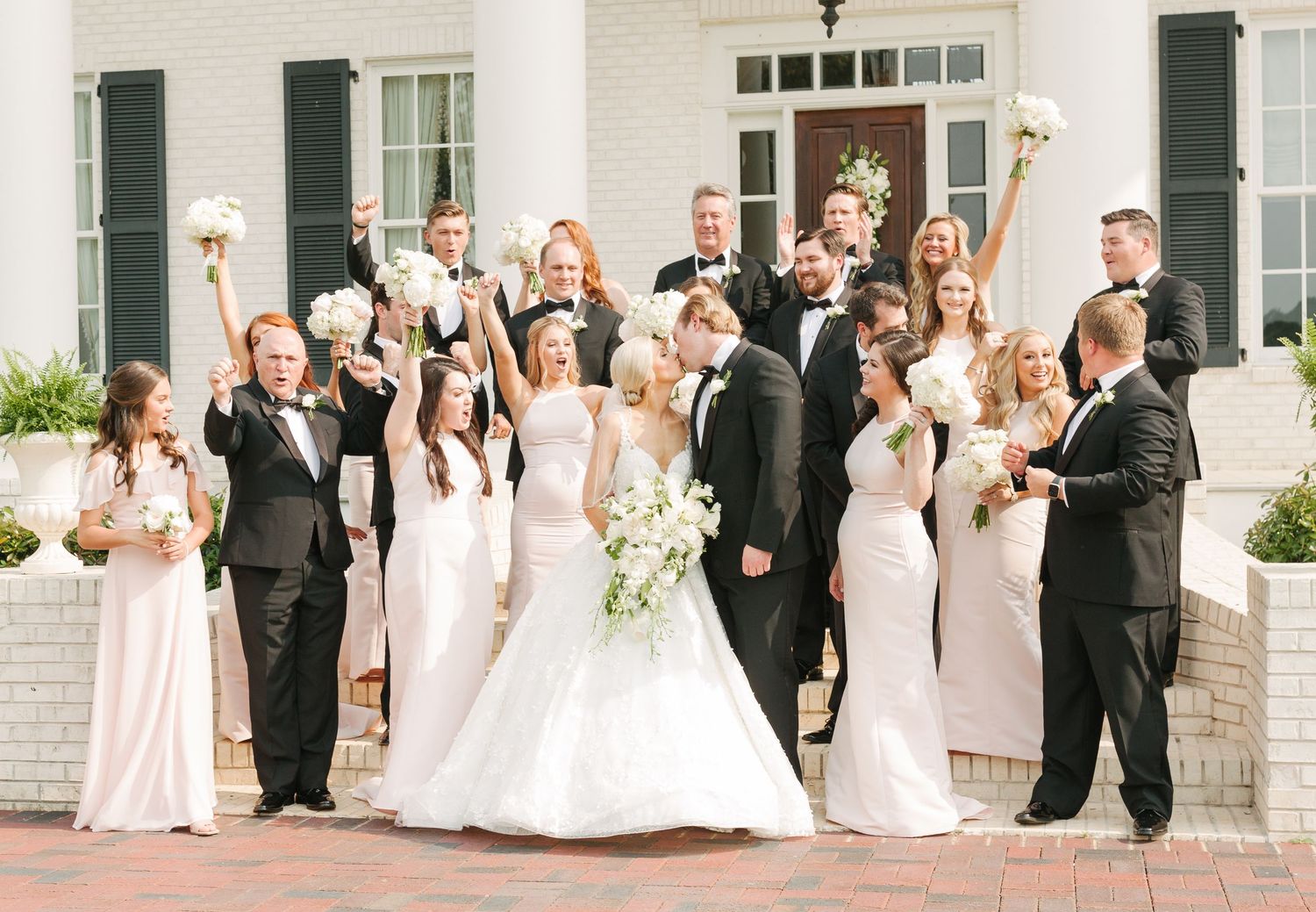 Wedding party in formal attire celebrates with raised arms and joyful expressions outside a white brick venue.