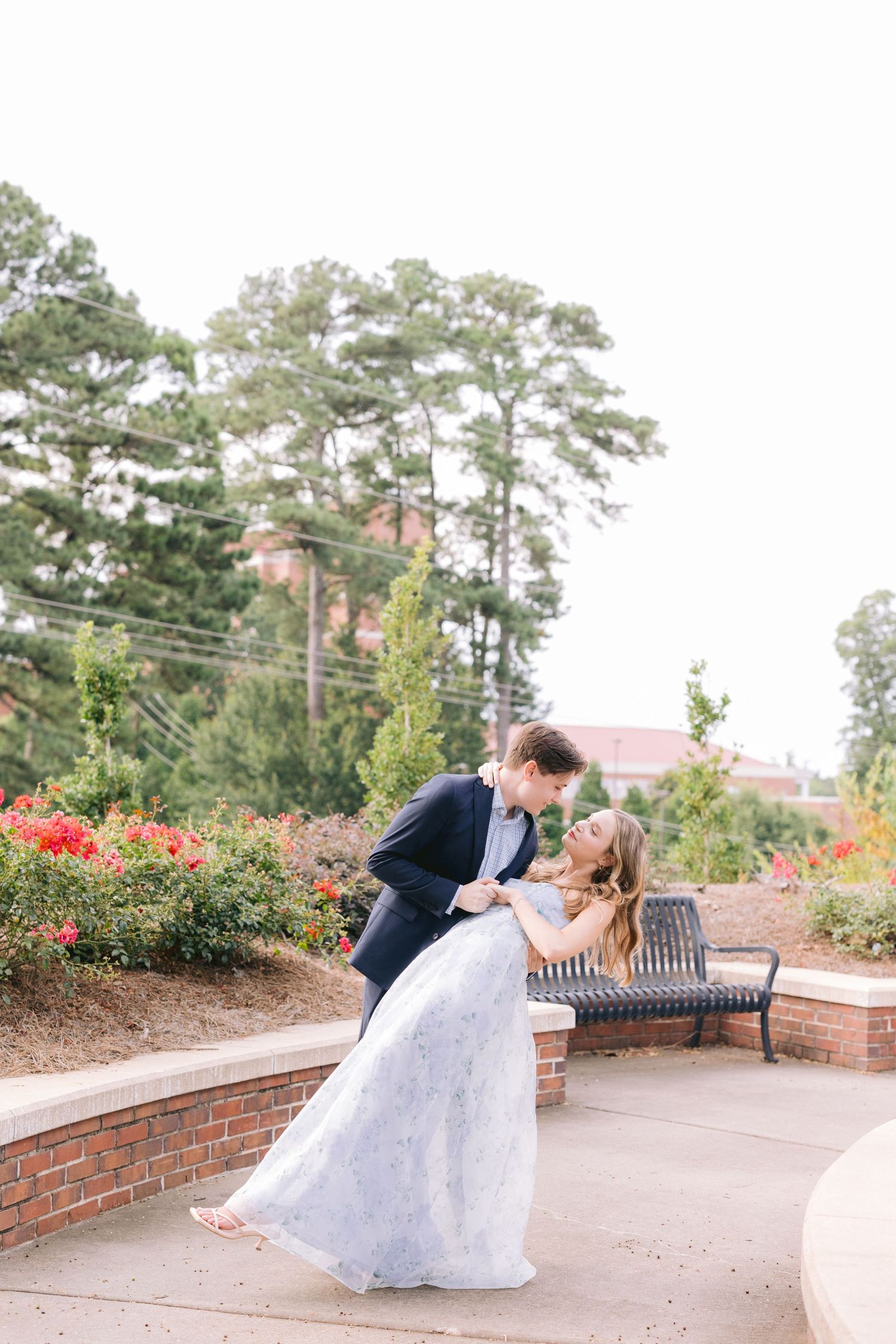 A romantic dip pose between newlyweds in front of red flowers and pine trees on a garden patio.