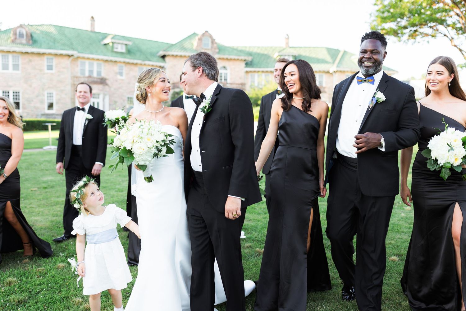 Wedding party laughs together outdoors in formal black and white attire on a sunny day.