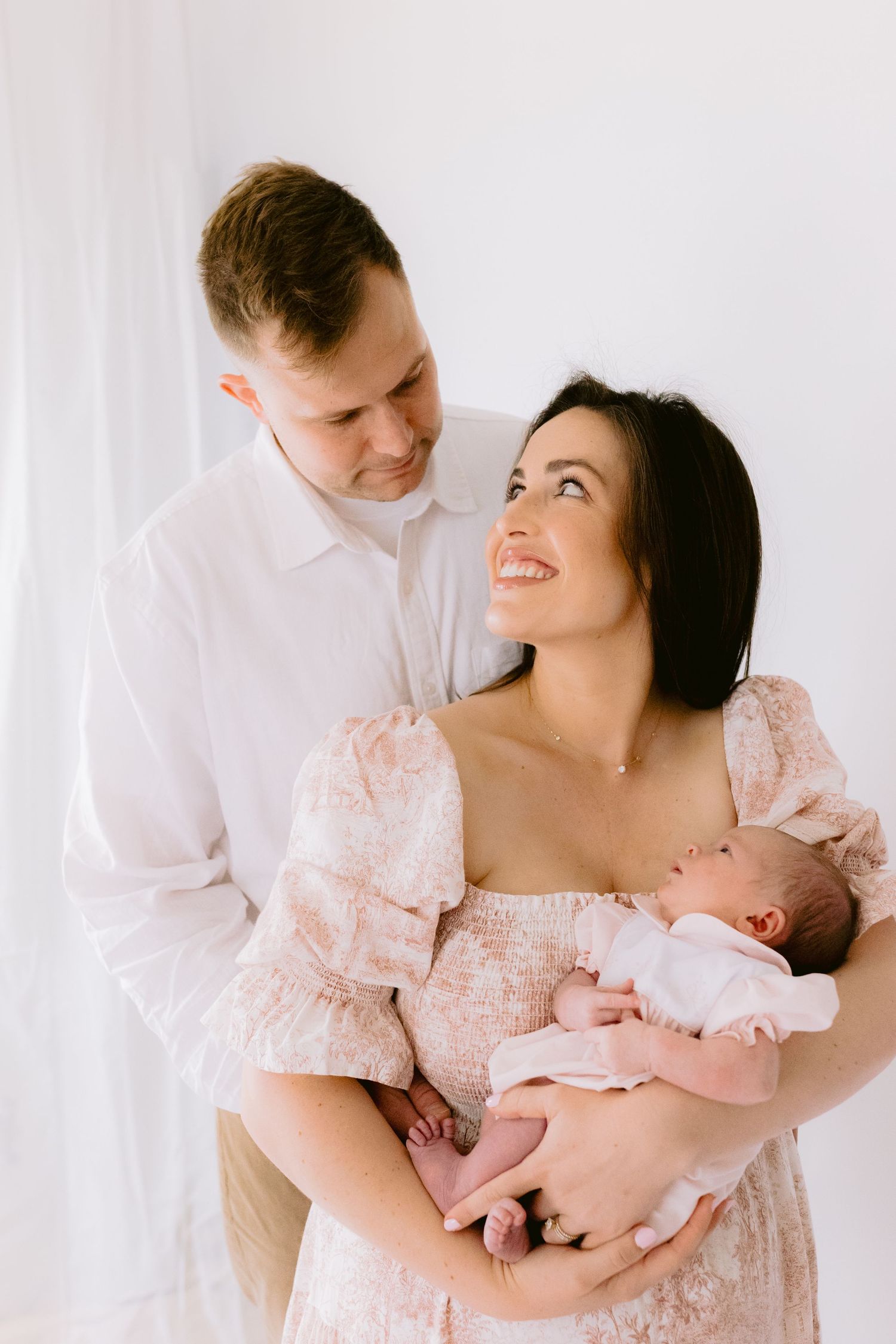 Happy couple in light clothing gazes lovingly at each other while holding their newborn baby against white backdrop.