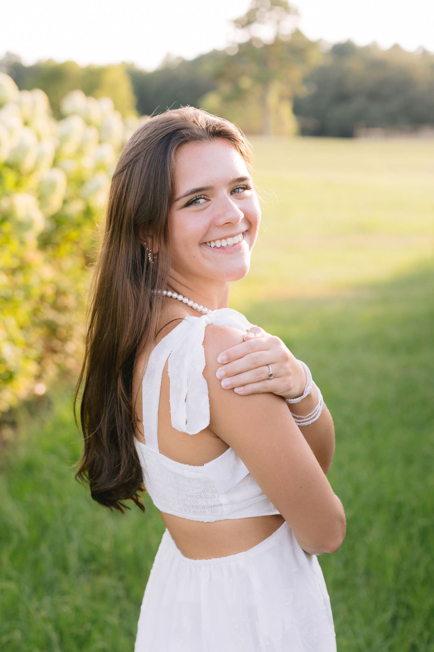 A person in a white dress poses outdoors in a sunny field during golden hour with soft natural lighting.