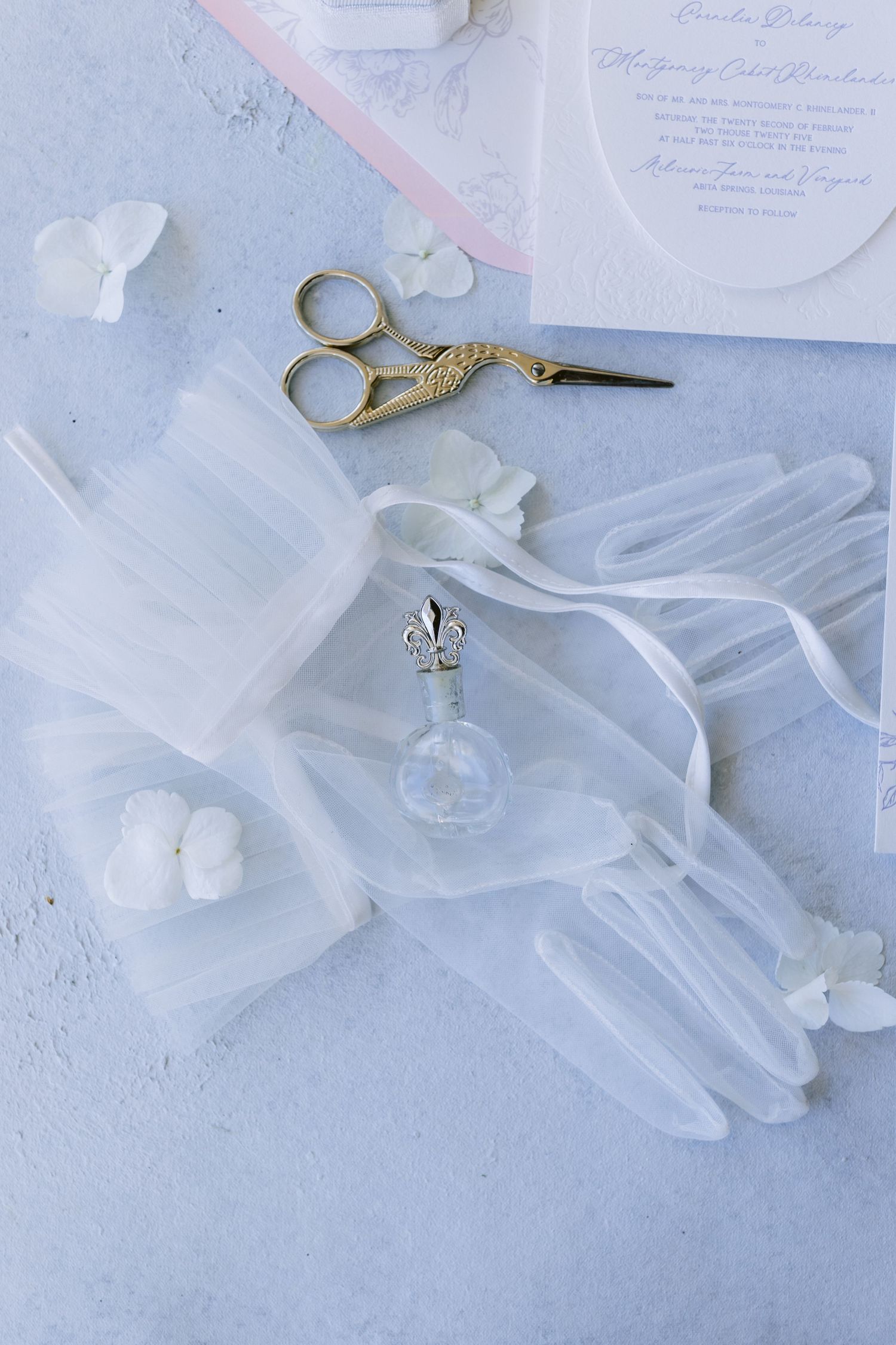 Wedding details flatlay featuring white gloves, gold scissors, and delicate invitations on light blue background.