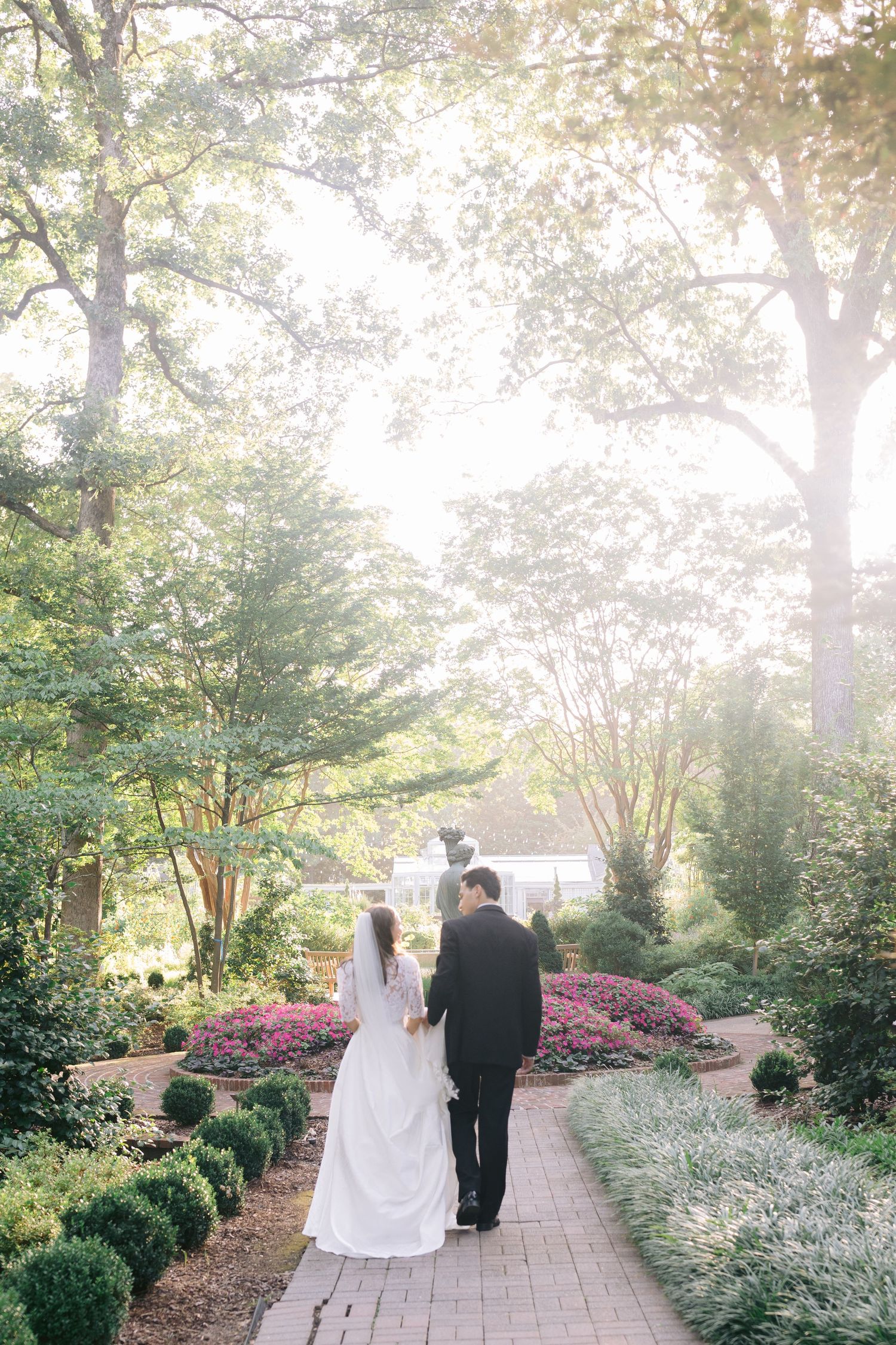 Couple walking down a garden path surrounded by blooming flowers and trees in golden sunlight.