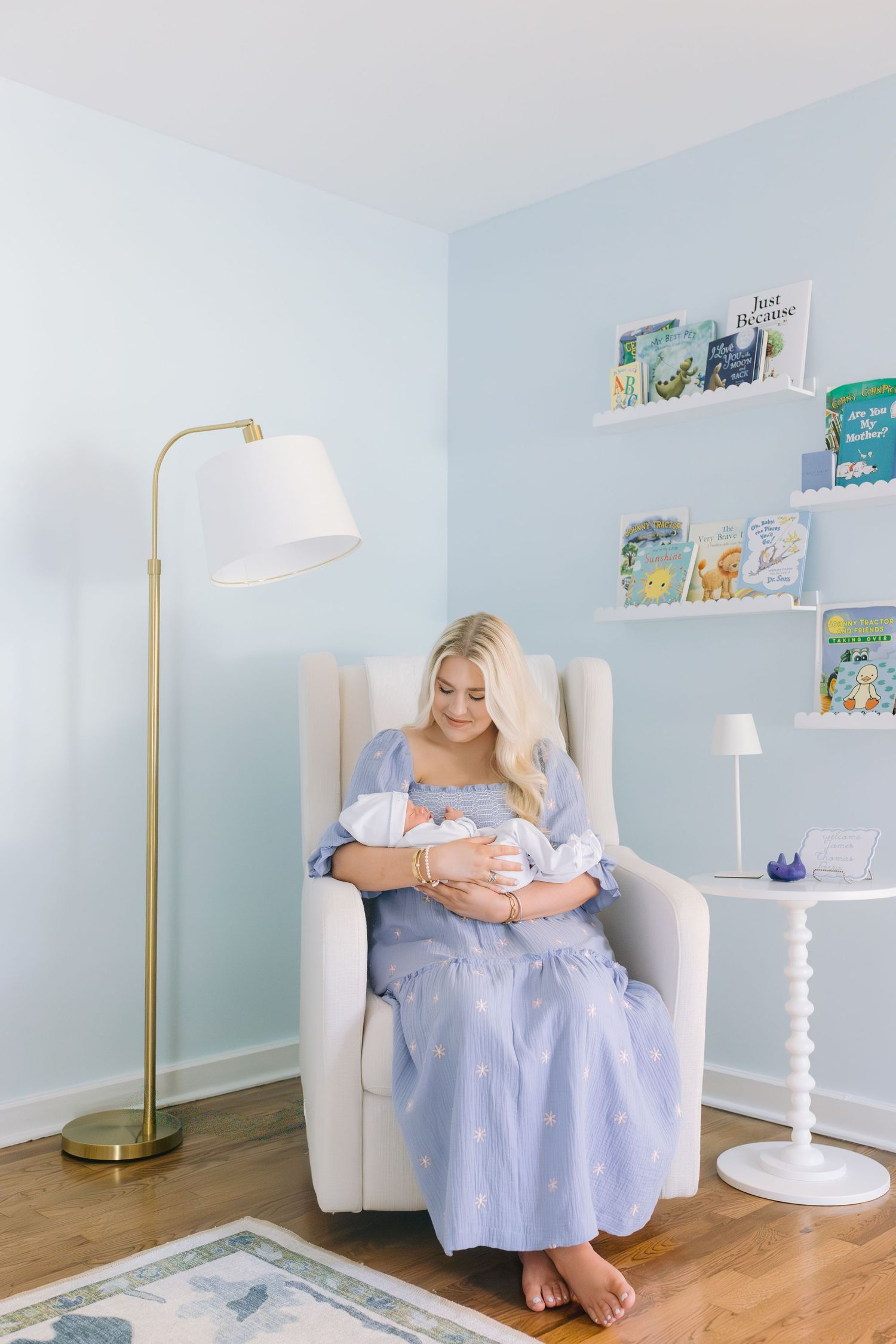 A light blue nursery room with white furniture and floating shelves displaying colorful children's books.