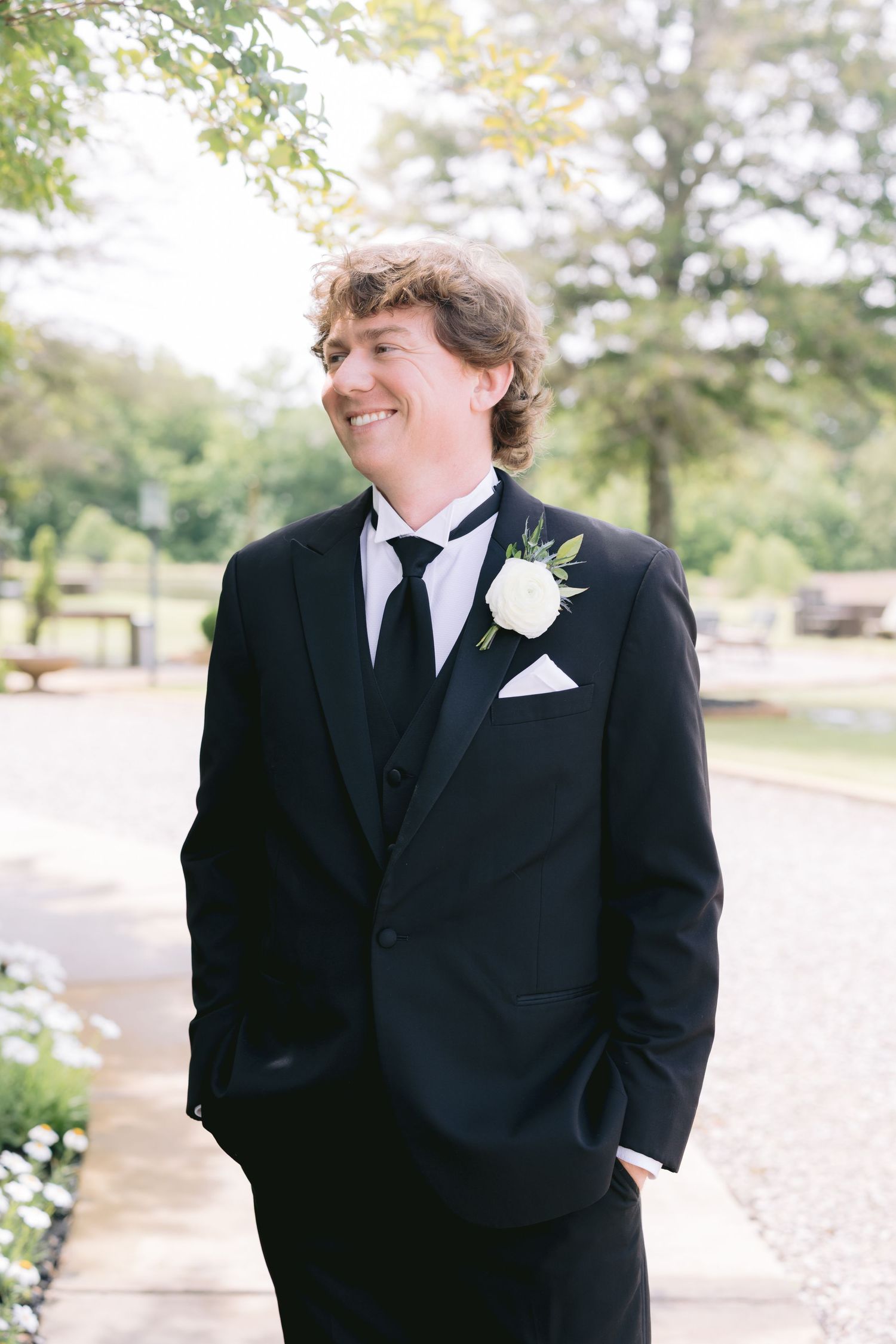 Wedding party member in black tuxedo with boutonniere standing outdoors.