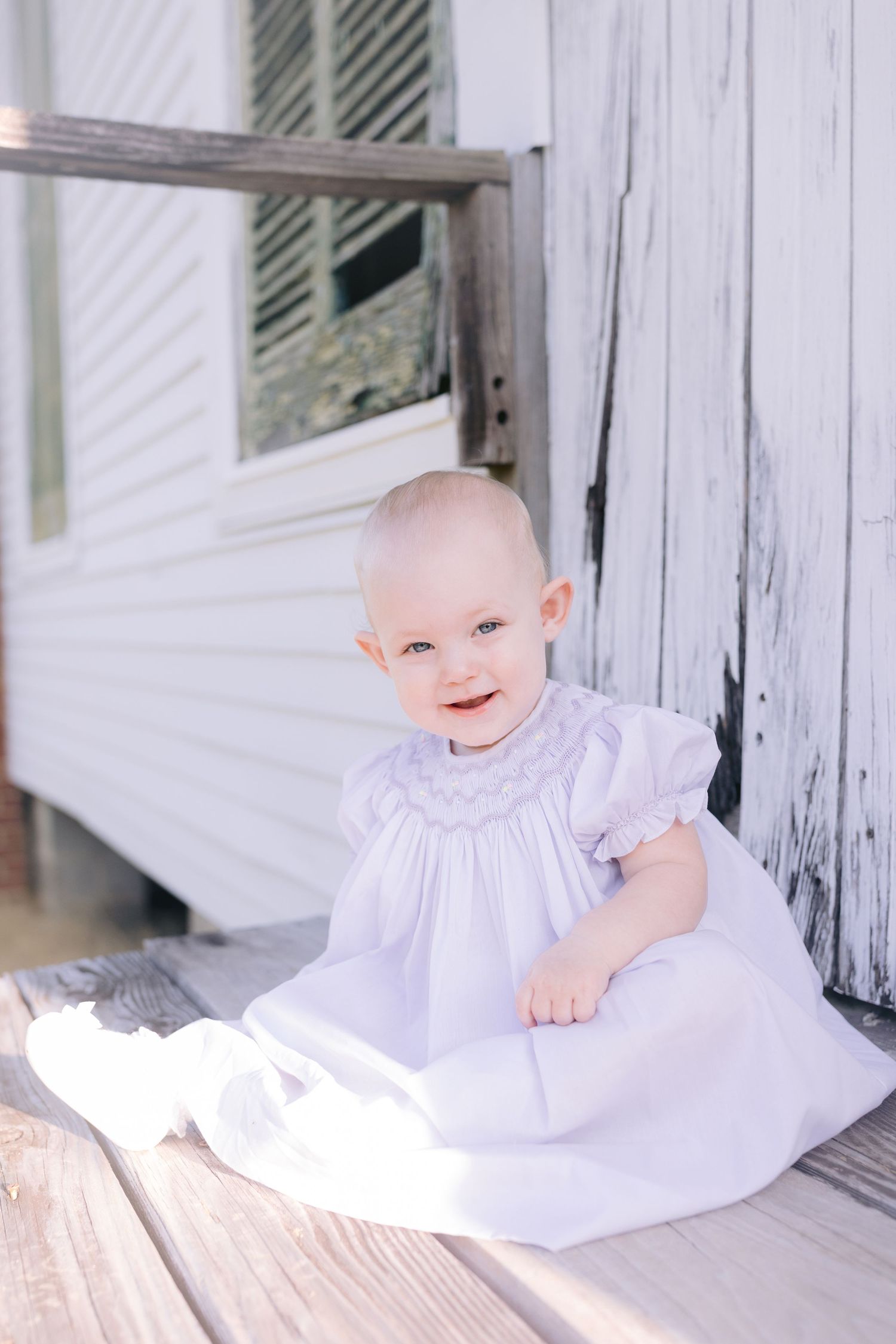 Sweet portraits of a baby in a white dress sitting against a weathered white wall.