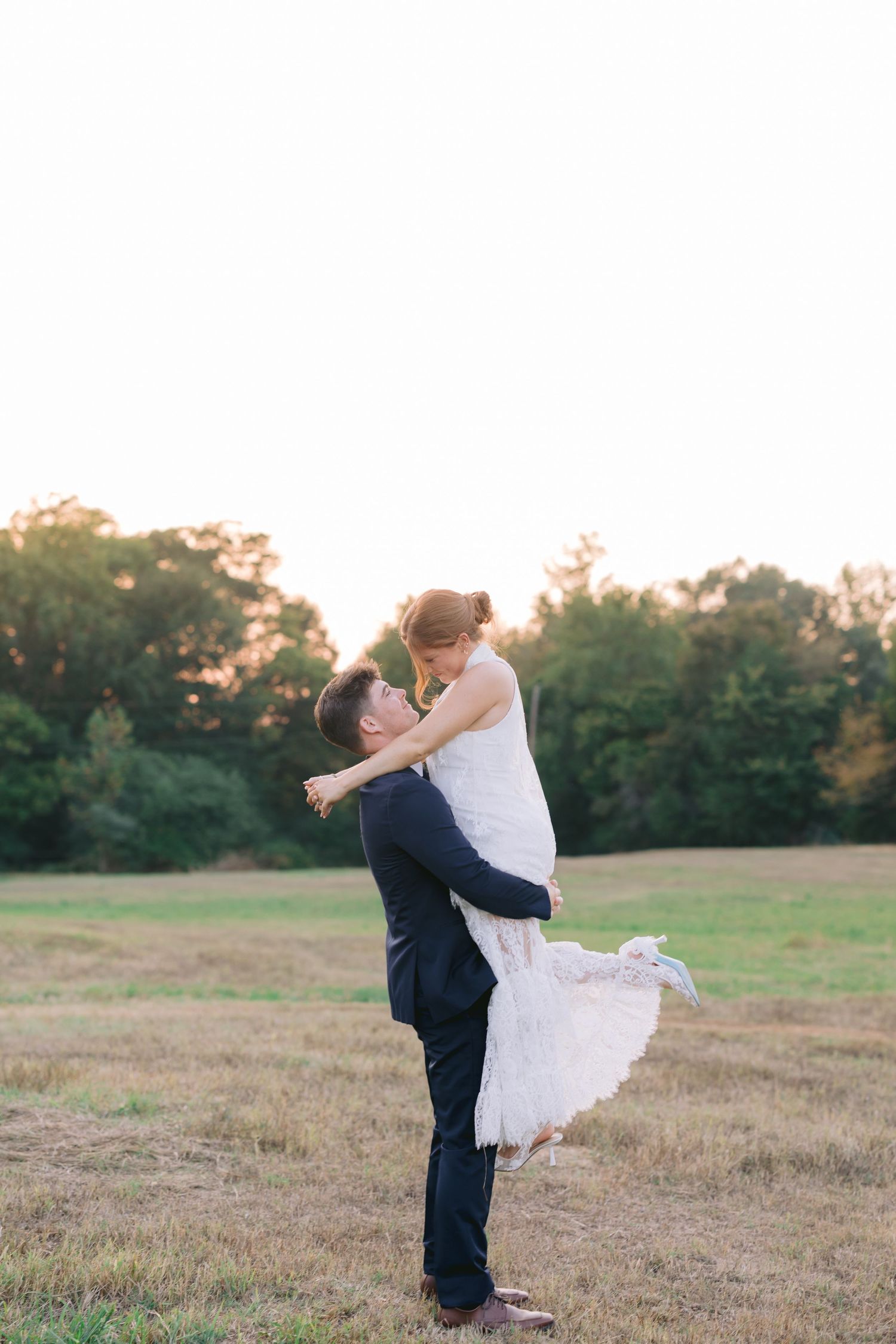 A couple shares a romantic kiss in a golden-lit field at sunset, with the groom lifting the bride in her white dress.
