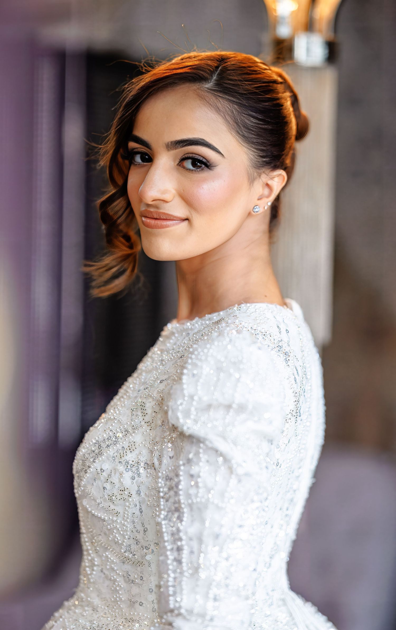A beautiful bride in a beaded white gown with an elegant updo smiles over her shoulder in a softly lit room.