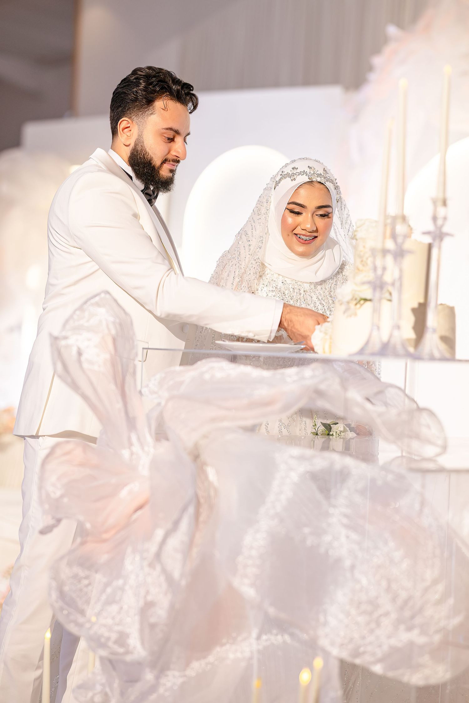 A wedding celebration scene with the couple in elegant white attire cutting their cake together.