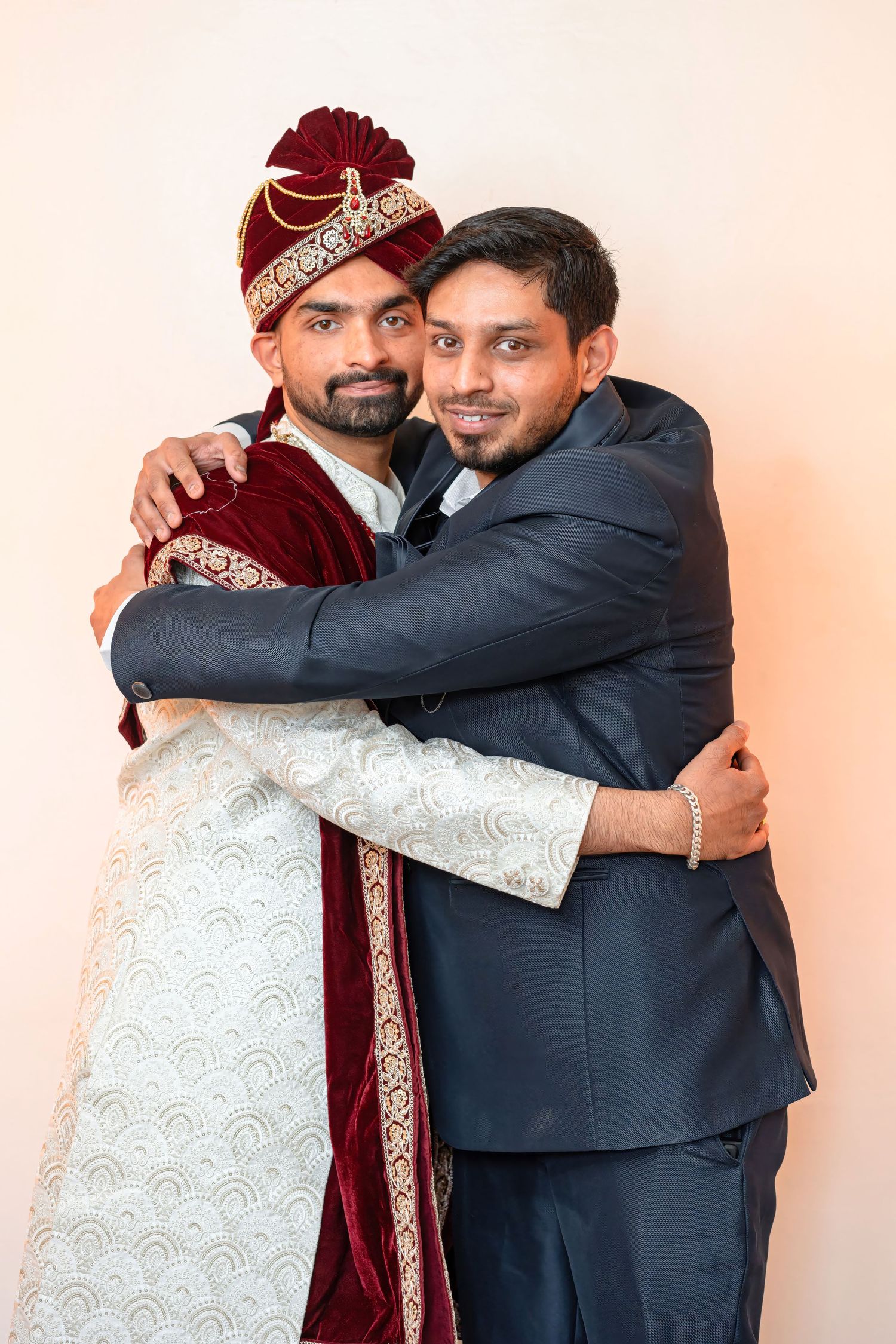 Two people embrace while one wears traditional South Asian wedding attire with a velvet sherwani and ornate turban.
