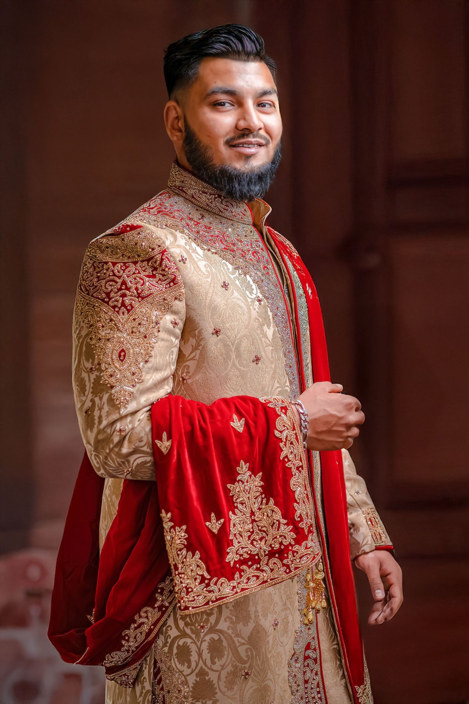 Person wearing an intricately embroidered cream and red sherwani with matching stole at a formal event.