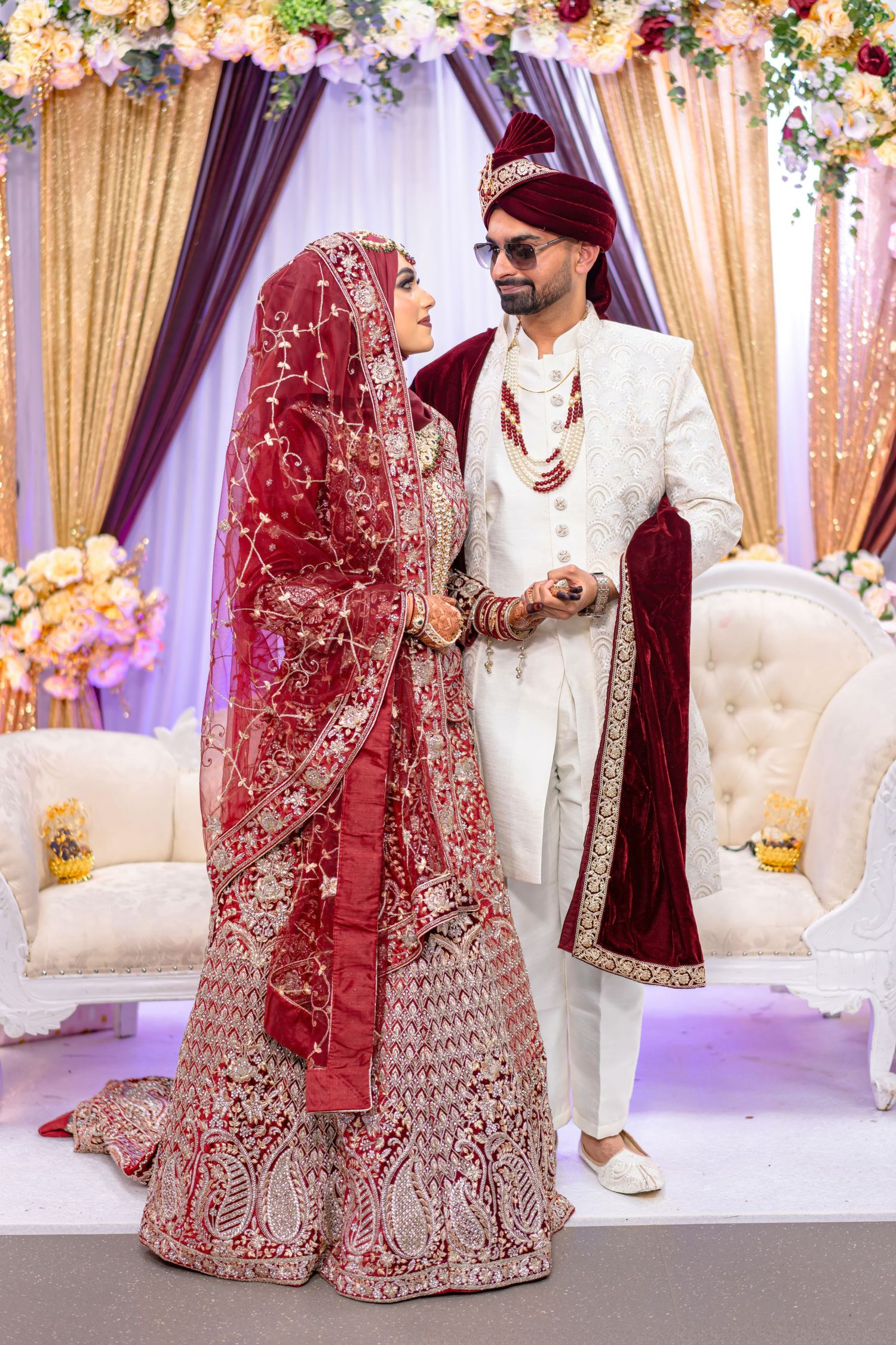 Newlyweds in red and white South Asian wedding attire posing under floral mandap decoration.