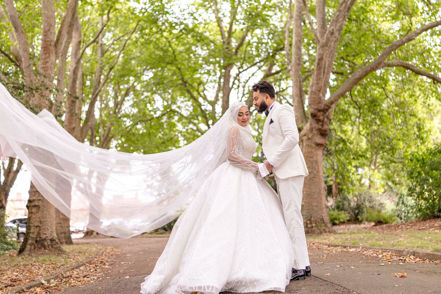 Wedding couple embraces on tree-lined path as bride's long veil flows dramatically in the breeze.