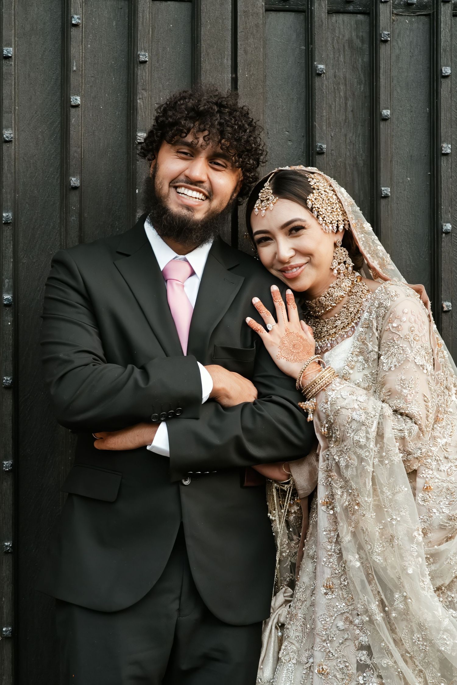 A smiling groom in a black suit with pink tie poses with his bride in a golden South Asian bridal outfit showing her ring.