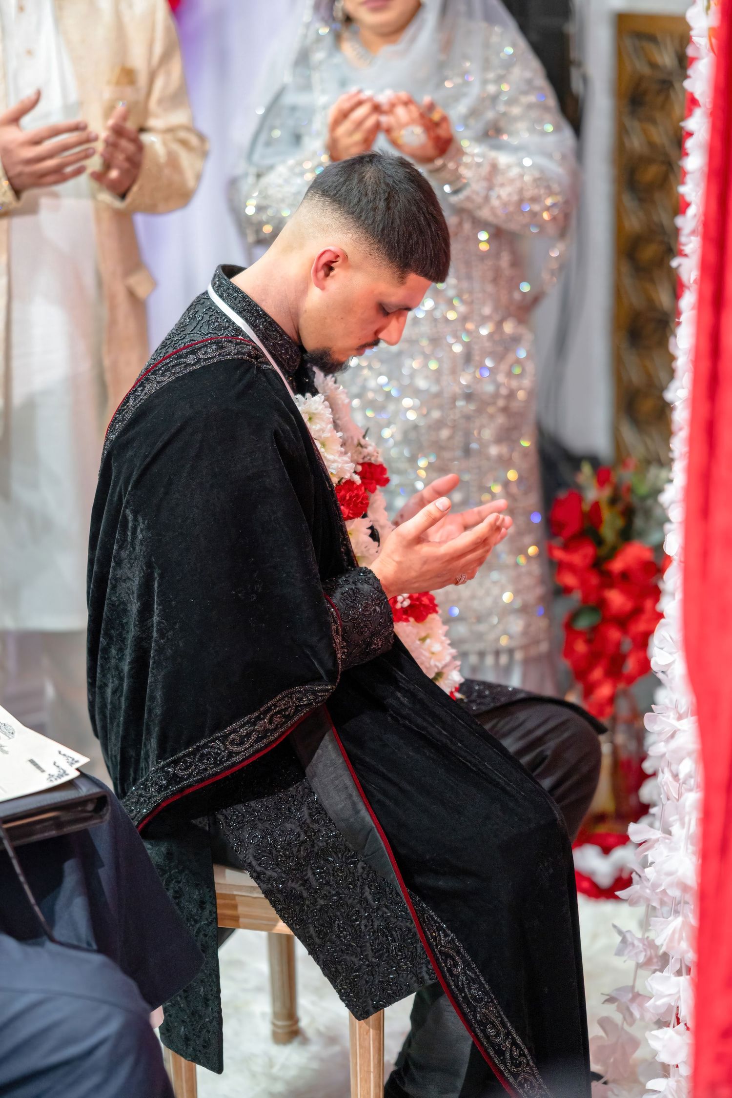Person in black formal attire praying during cultural ceremony surrounded by red and white decorations.