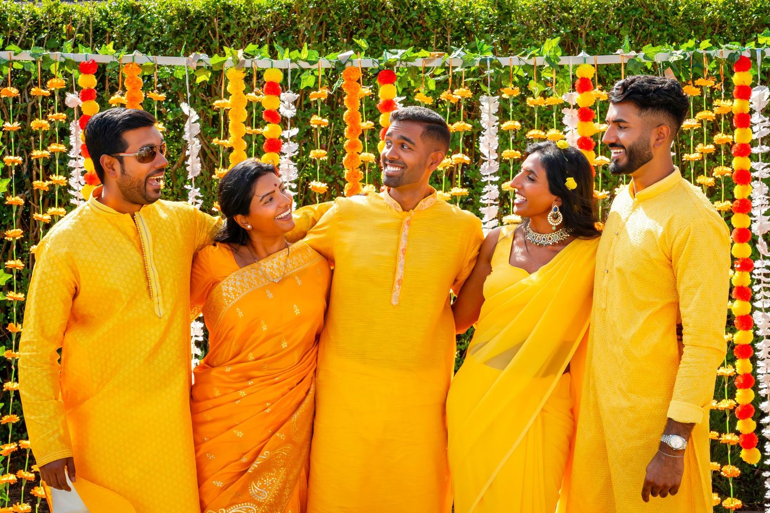 Five people in matching yellow traditional outfits pose against a floral backdrop.