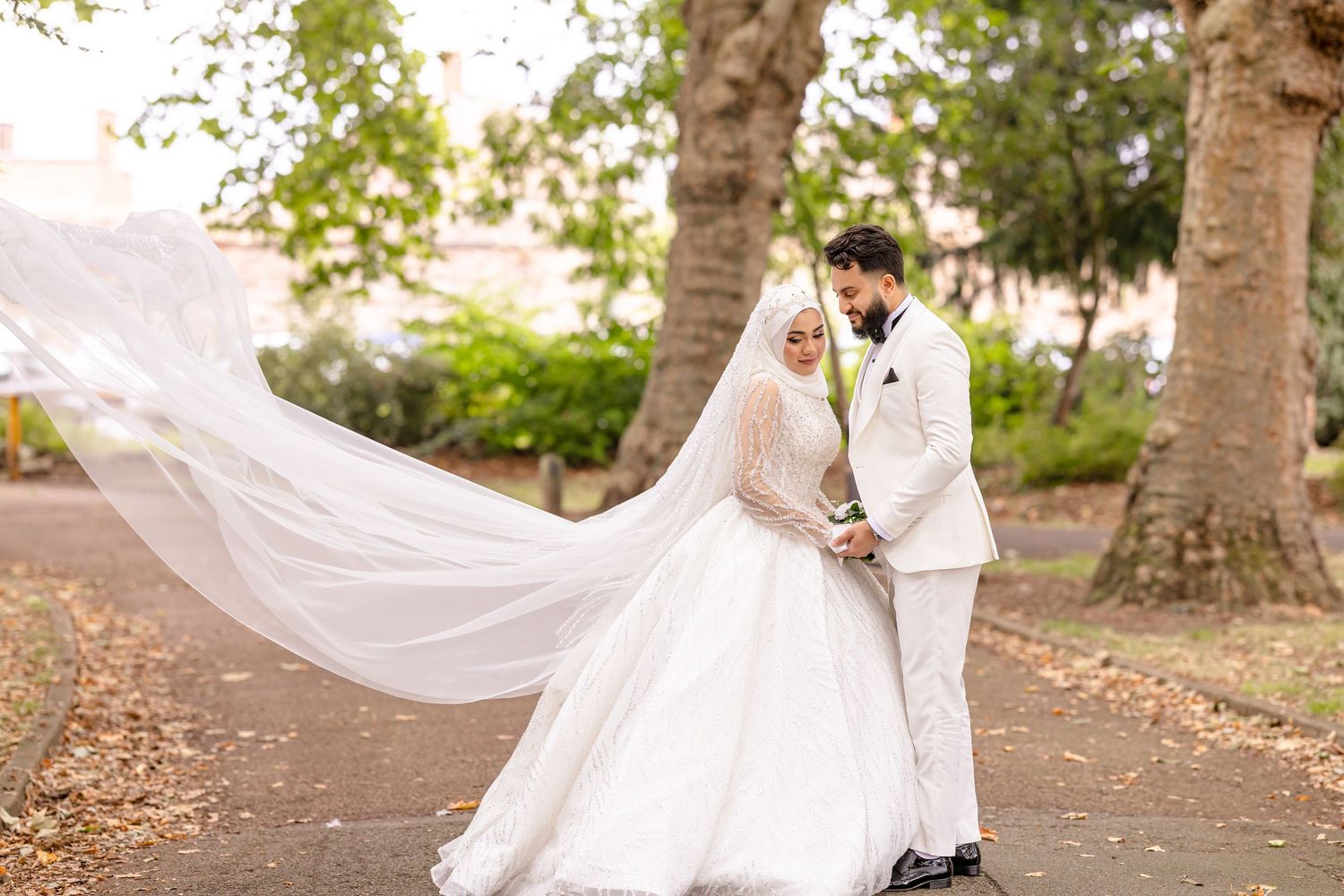 A couple shares a romantic moment on their wedding day in a sunlit park path with a flowing white veil.