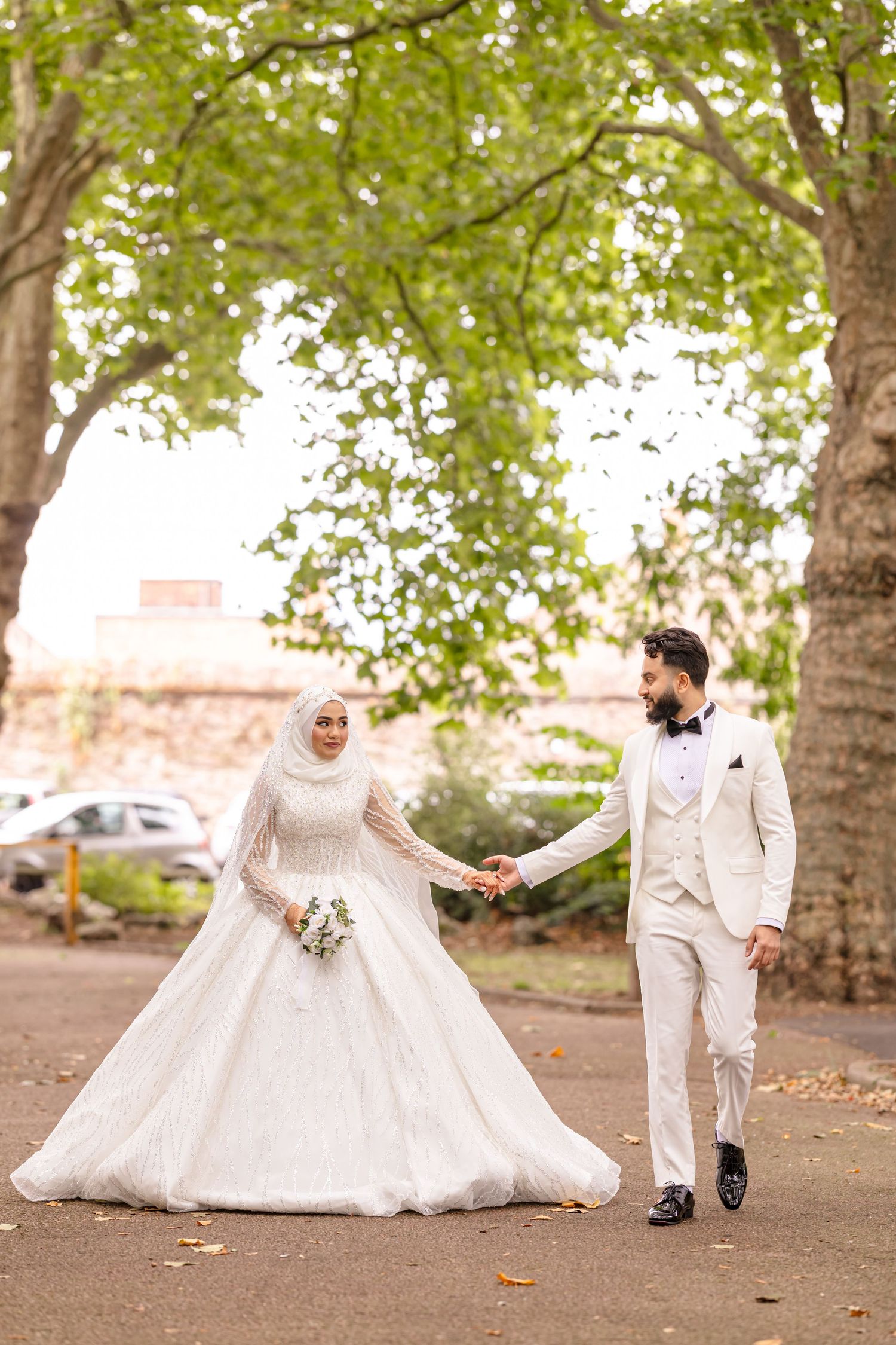 A bride in white hijab dress and groom in light suit hold hands while walking beneath green trees in an outdoor setting.